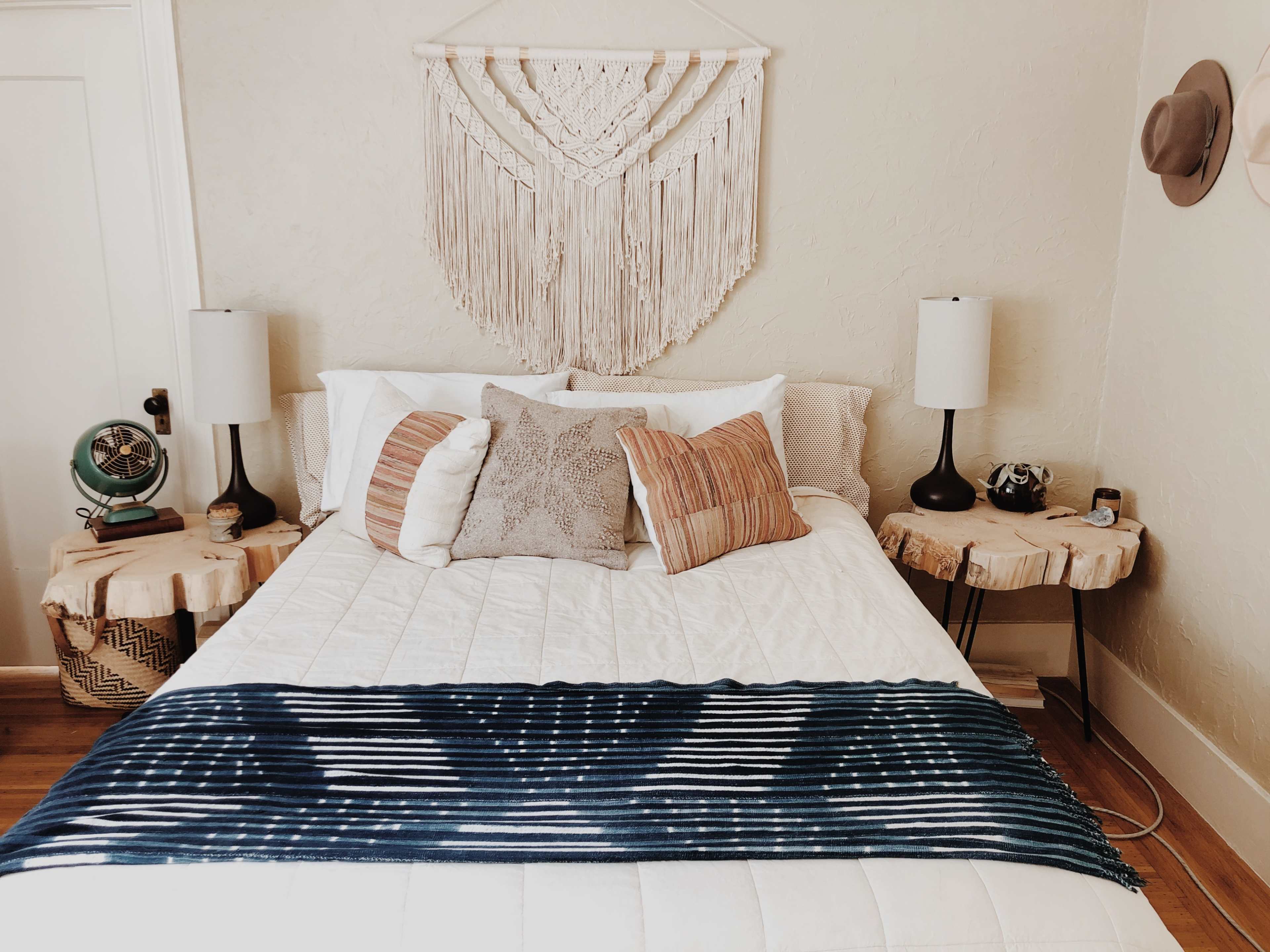 A neatly arranged bedroom features a bed with decorative pillows, a macramé wall hanging, and wooden side tables with lamps.