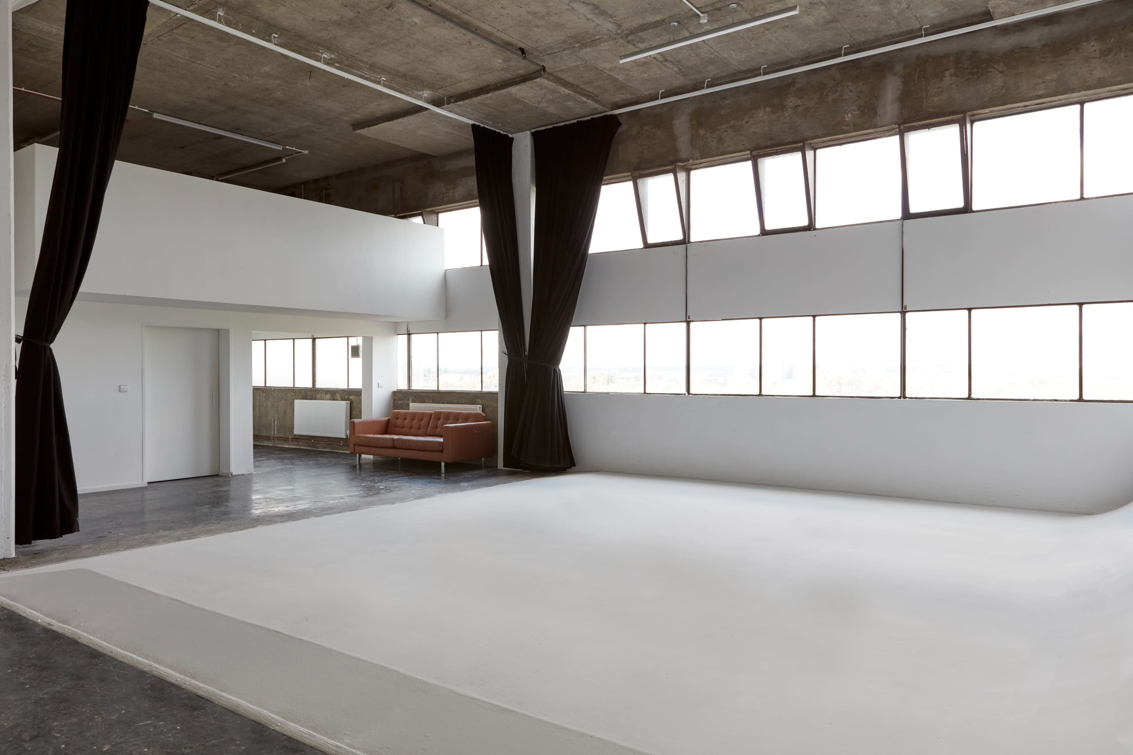 A spacious, minimalist warehouse-style interior with large windows, a white floor area, and a brown sofa against the wall.