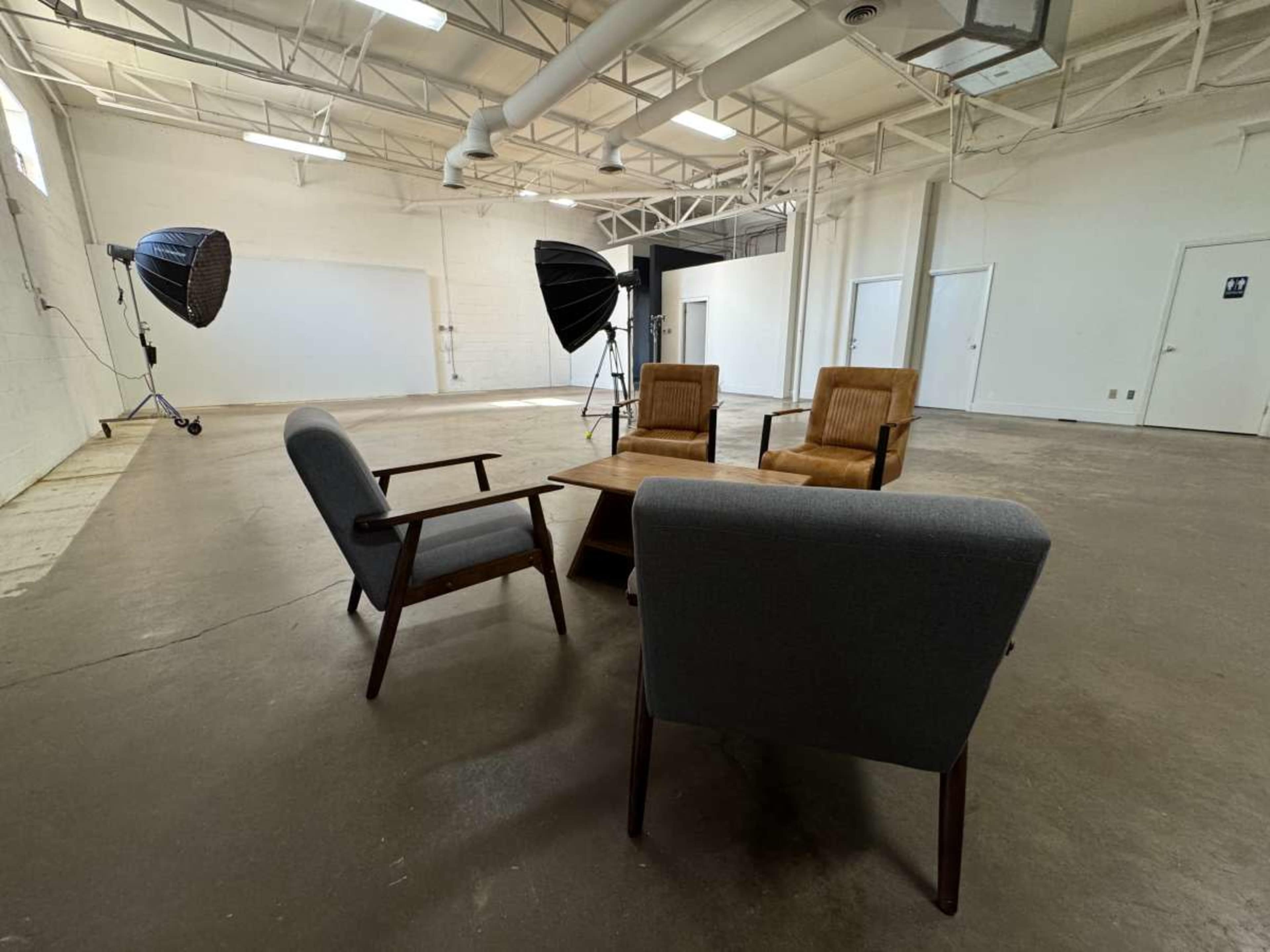 The image shows a spacious, well-lit room featuring two brown chairs and one gray chair arranged around a wooden table, with large studio lights positioned in the background.