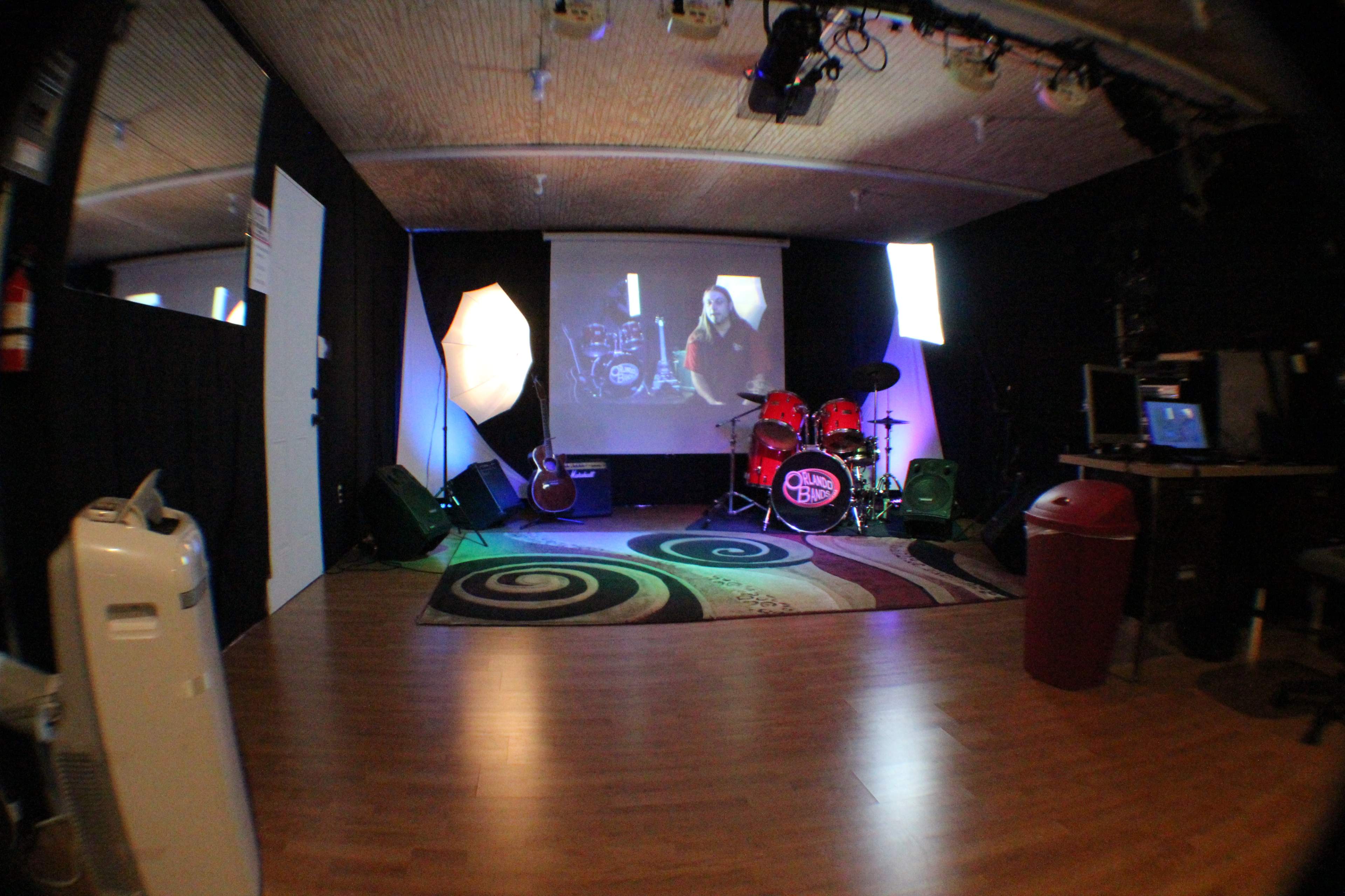 The image shows a music rehearsal space with a drum set, guitar, and various lighting setups, along with a computer and a video display.