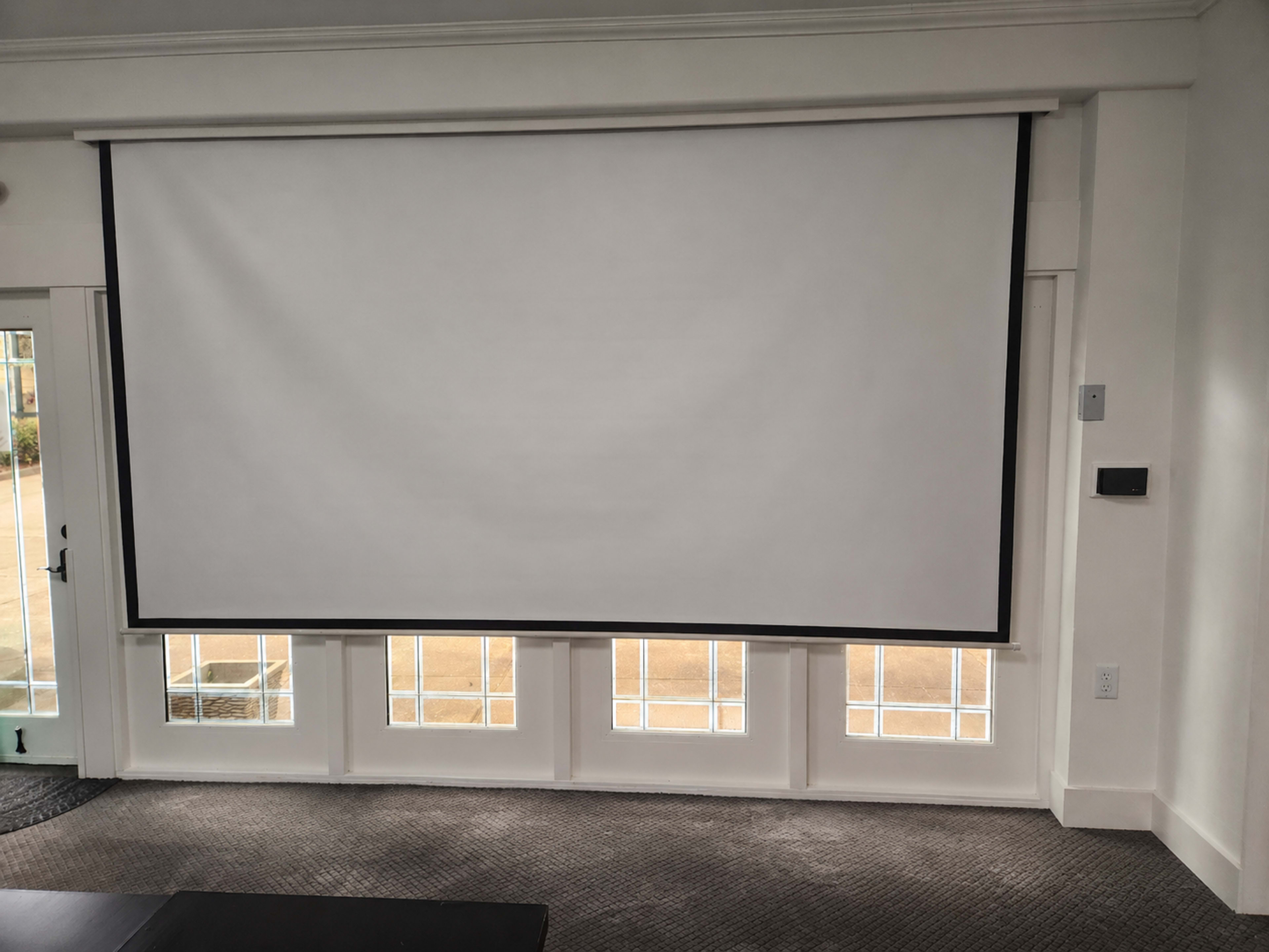 A large white projection screen is mounted on a wall above a row of rectangular windows with clear glass panes.