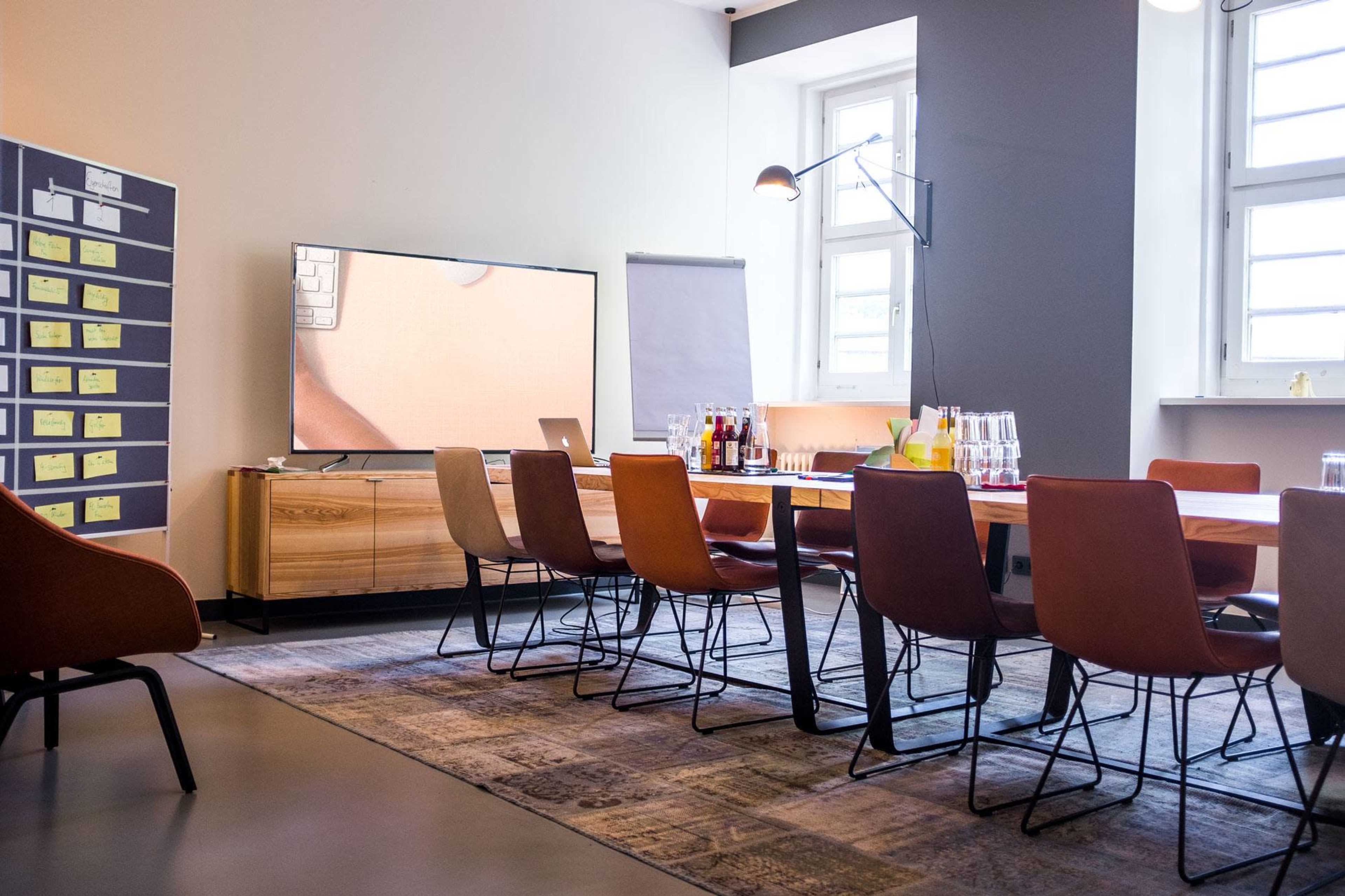 A conference room features a long wooden table surrounded by chairs, with a large television screen and a flip chart in the background.