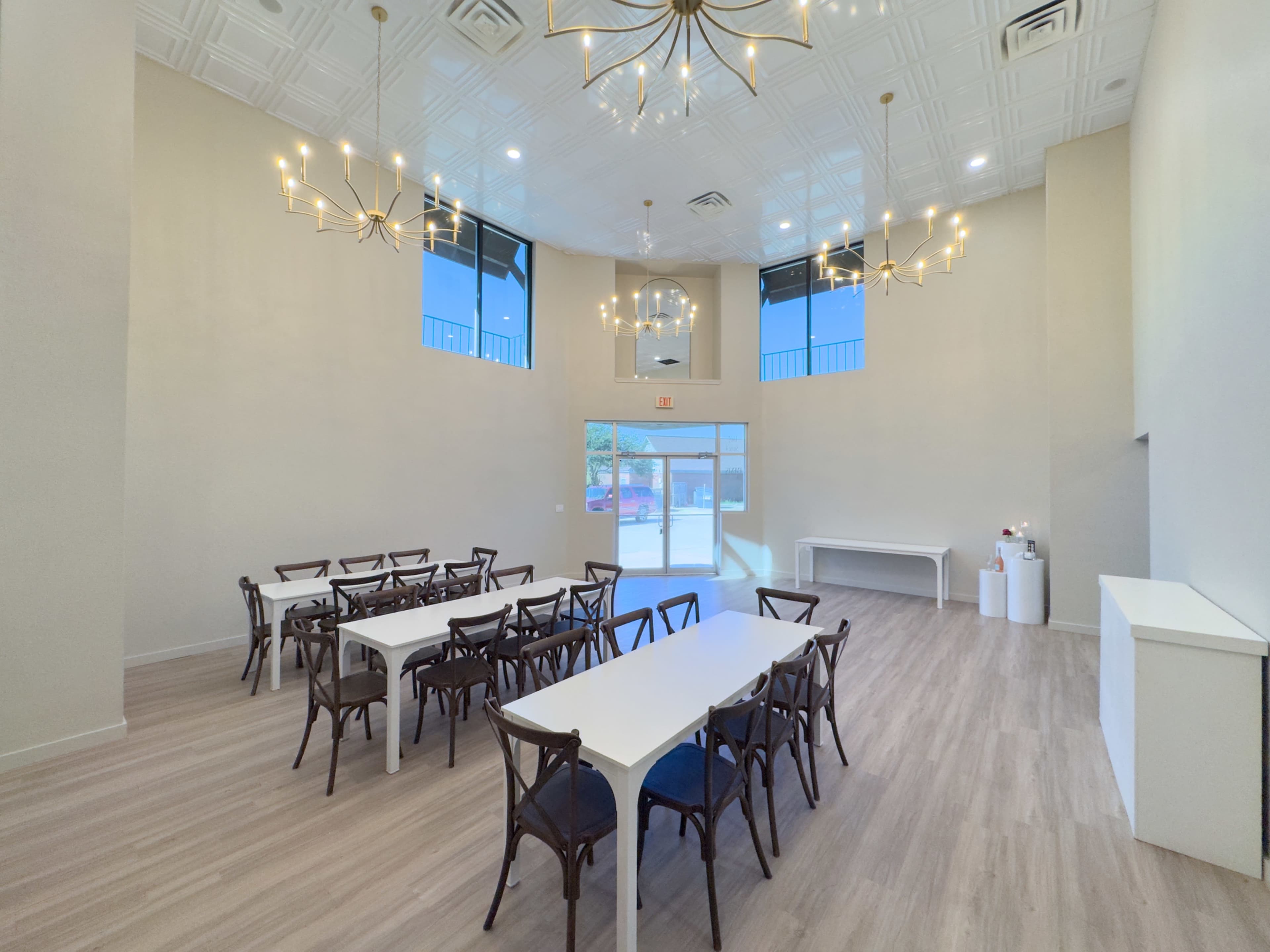 The image shows a spacious meeting room with multiple white tables and chairs, large windows allowing natural light, and decorative light fixtures hanging from the ceiling.