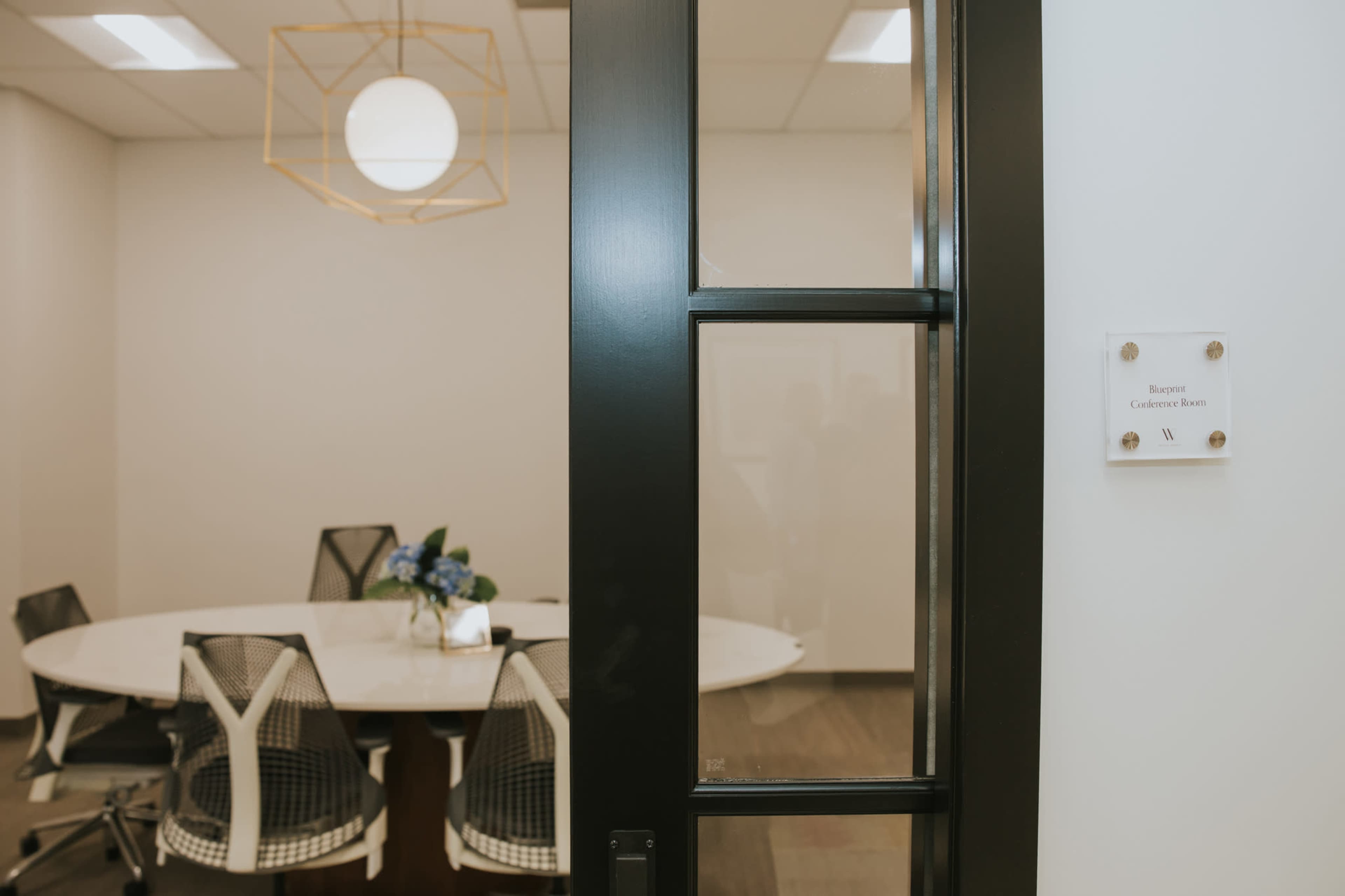 The image shows a modern conference room with a round table surrounded by mesh office chairs, viewed through a glass door with a sign identifying the room.