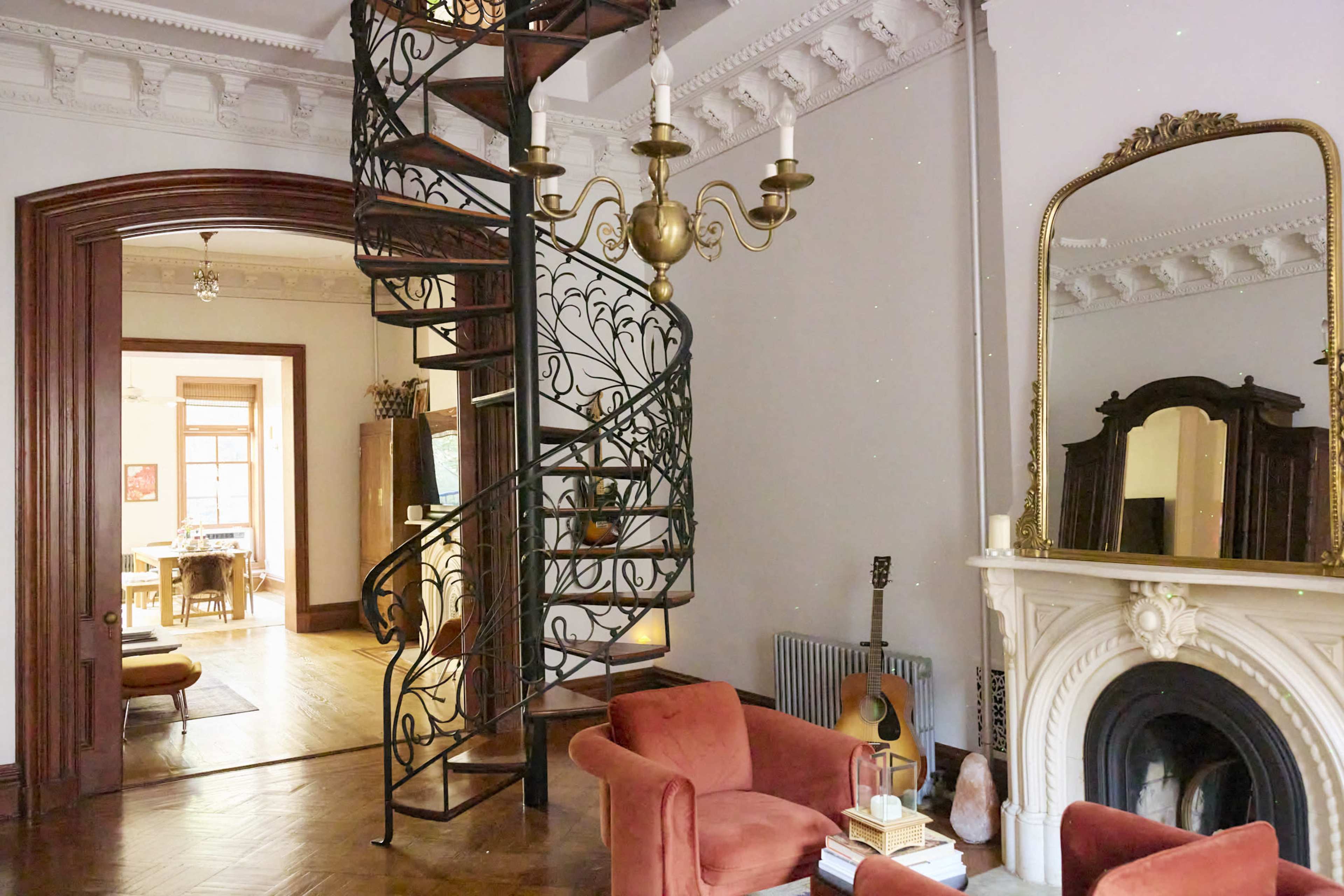A spiral staircase with decorative railings ascends beside a cozy living area furnished with orange chairs and a marble fireplace.