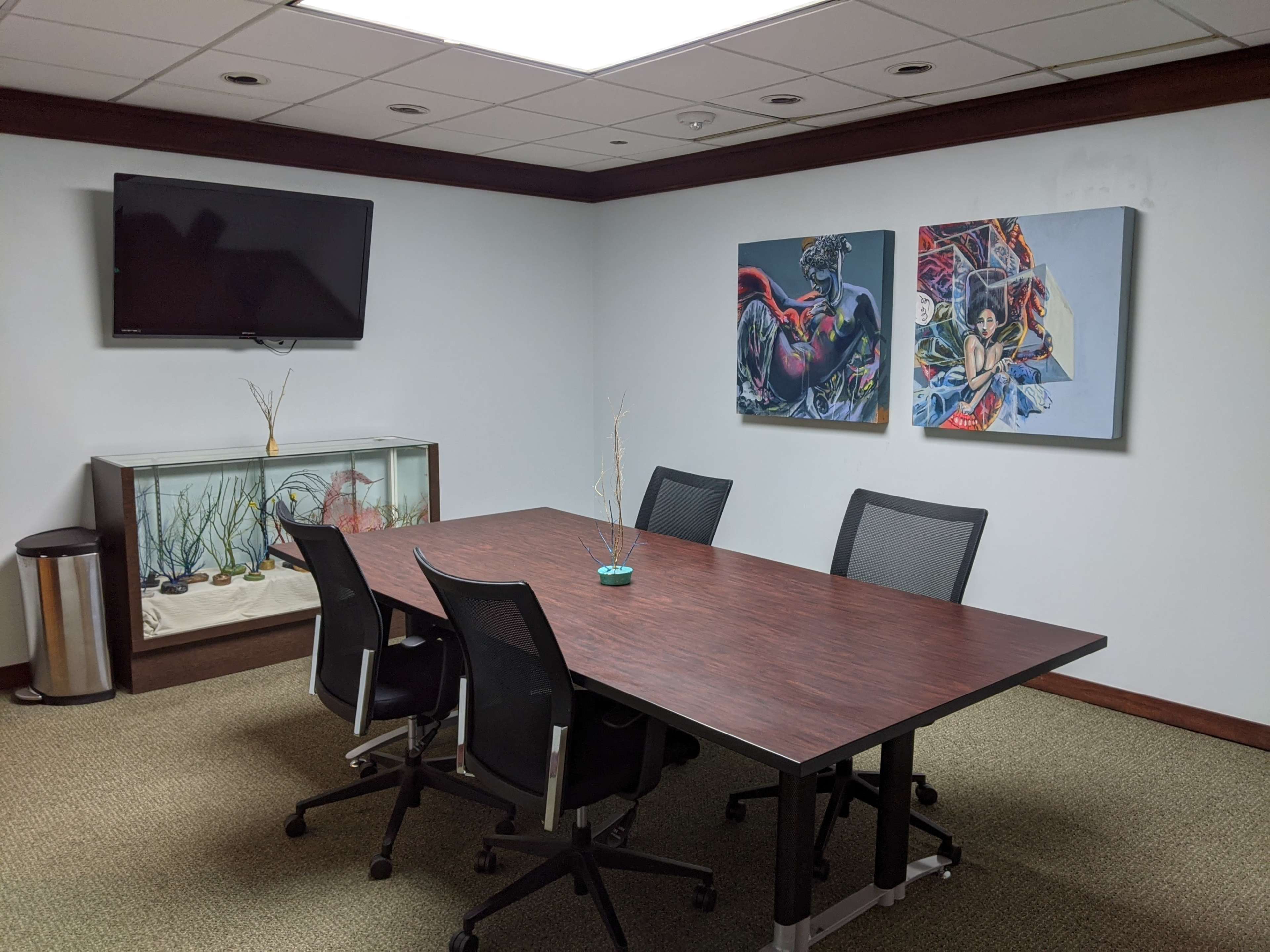 The image shows a small conference room featuring a large wooden table surrounded by black chairs, a wall-mounted TV, and two colorful paintings, along with an aquarium in one corner.