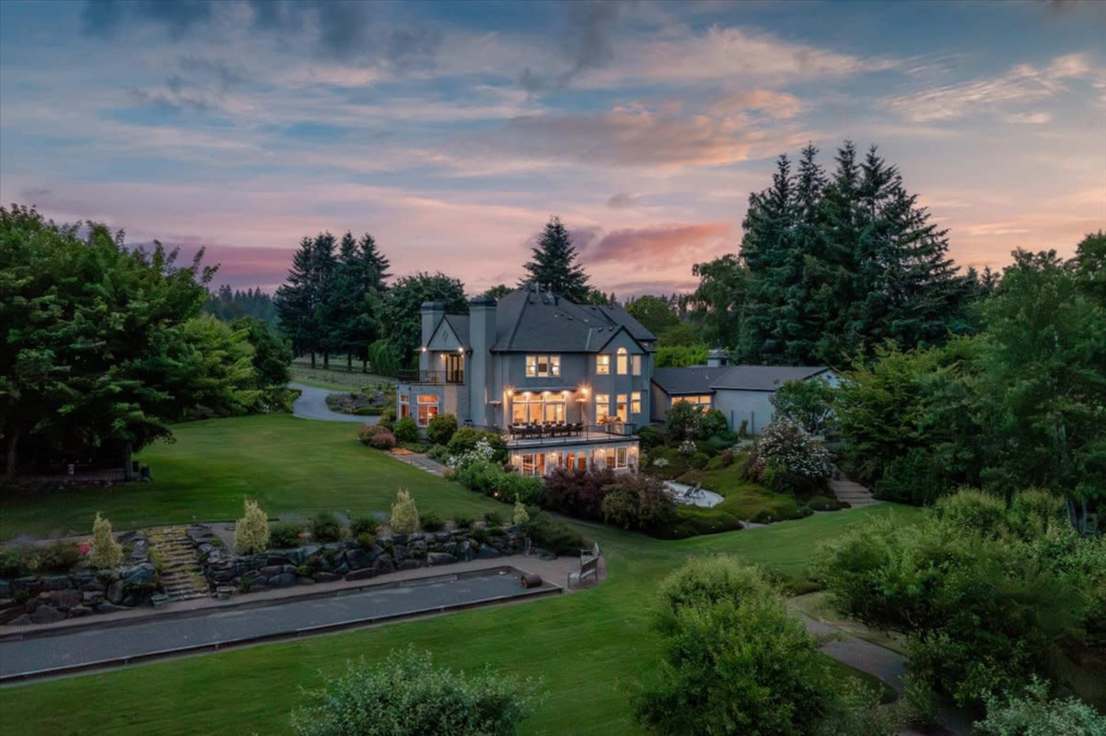 A large house with illuminated windows is surrounded by lush greenery and a well-maintained lawn at dusk.