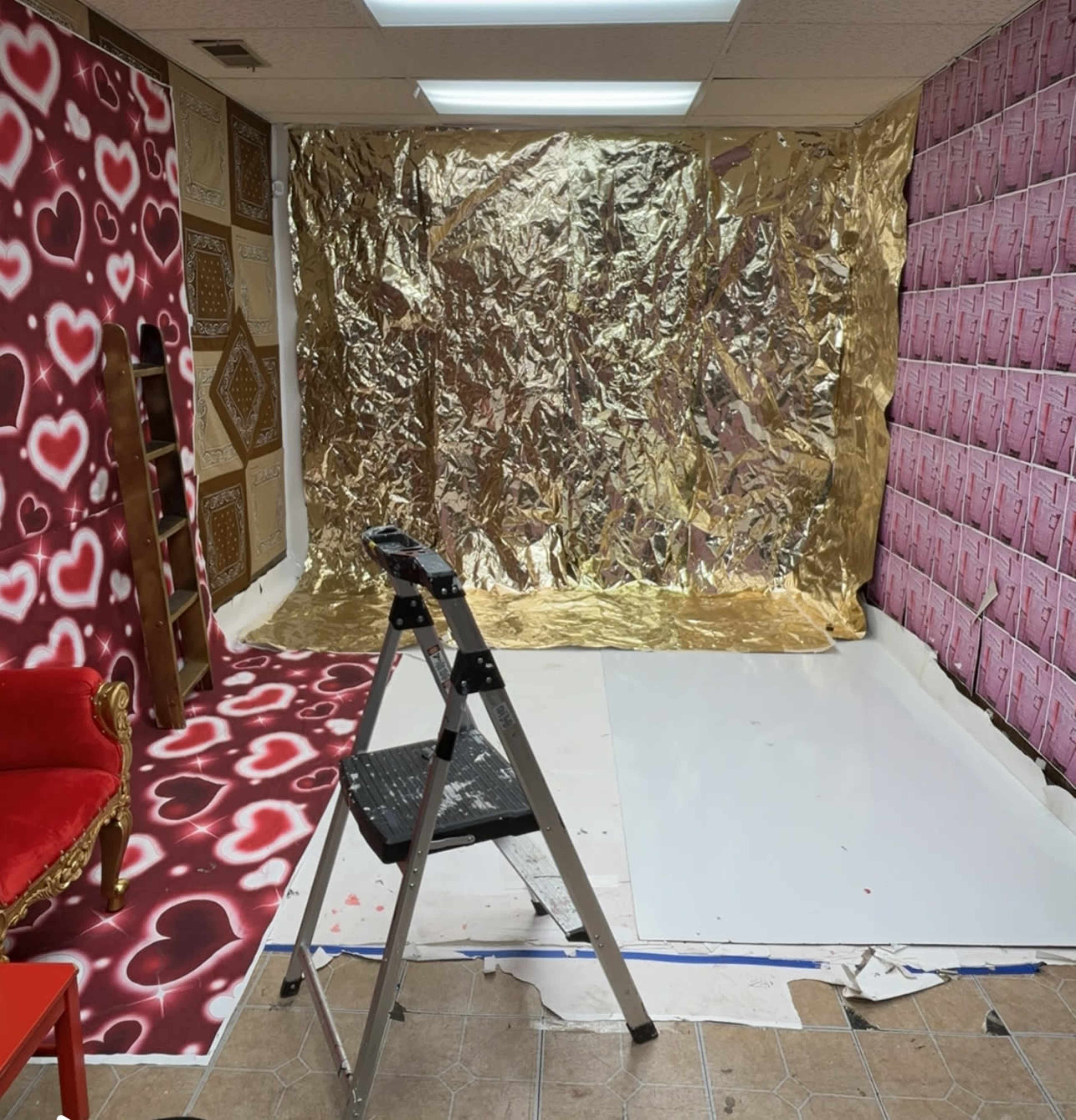 The image shows a room with a gold foil backdrop, heart-patterned walls, a white floor covering, and a ladder positioned on an unfinished floor.