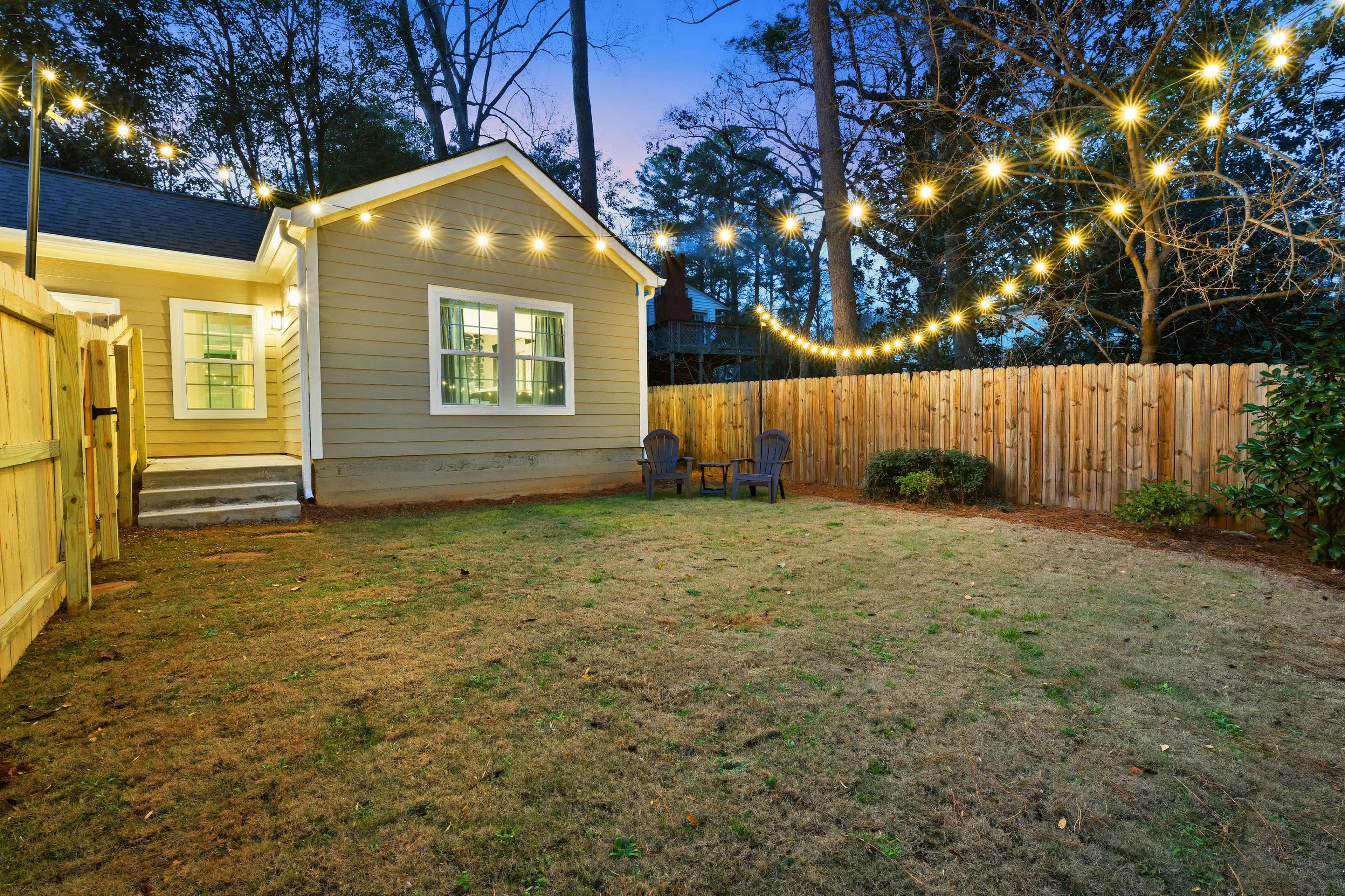 A backyard features a small patch of grass, two chairs arranged beside a decorative light string hanging between trees, with a wooden fence enclosing the space.