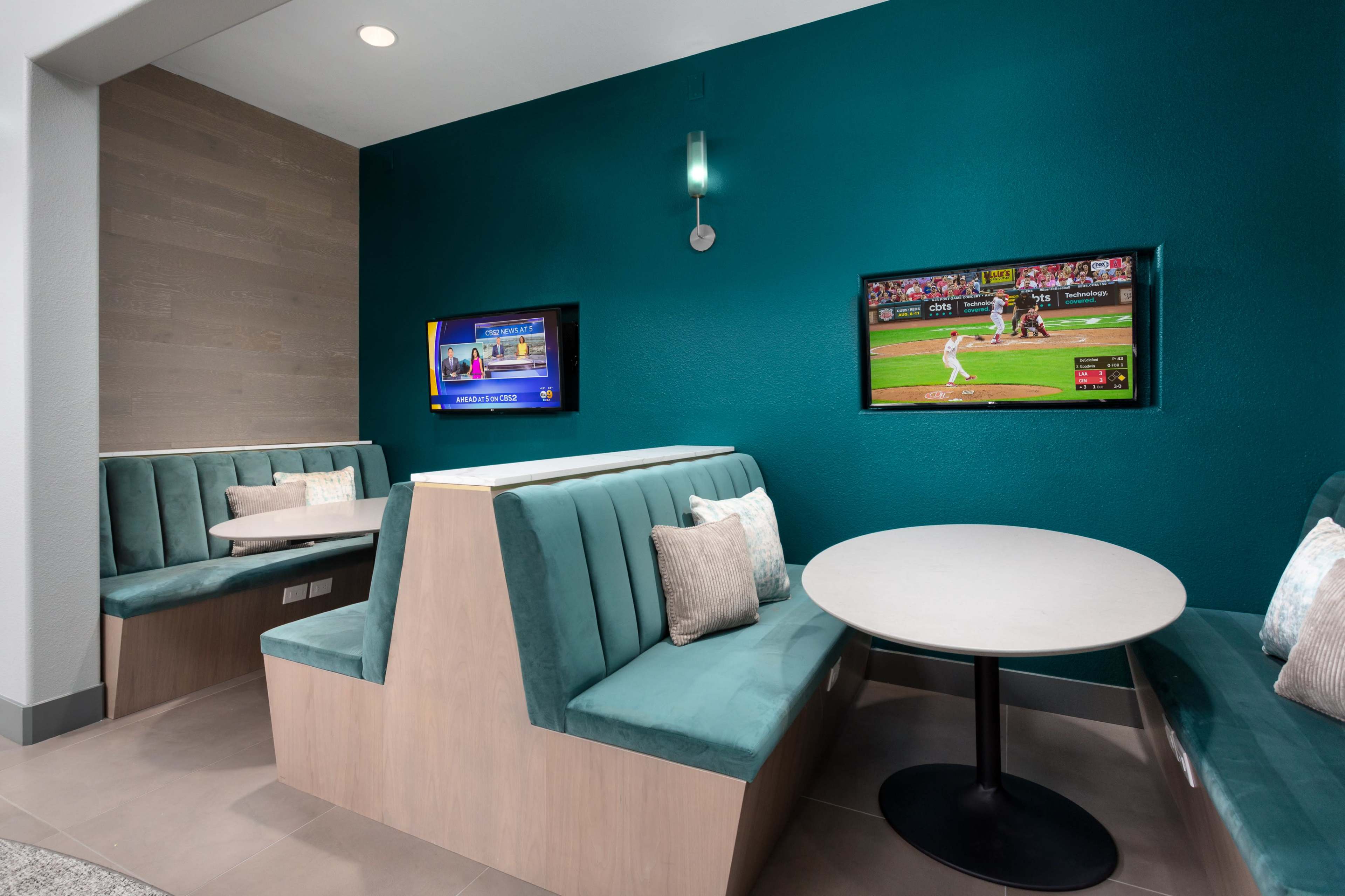 The image shows a cozy seating area with teal-colored walls, featuring two television screens and a round table surrounded by cushioned booths.