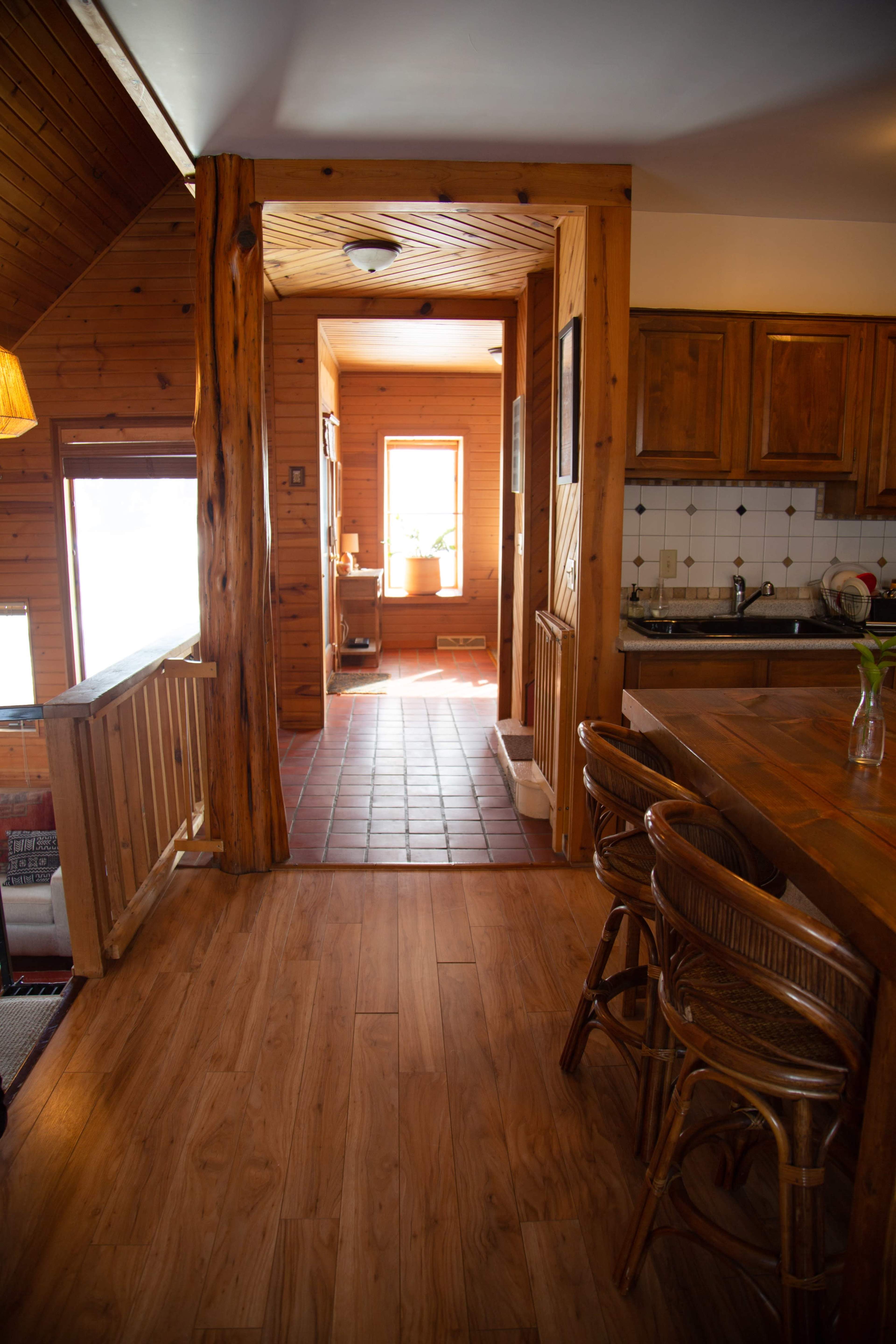A wooden interior features a kitchen area with bar stools and a view of a hallway leading to a well-lit room.
