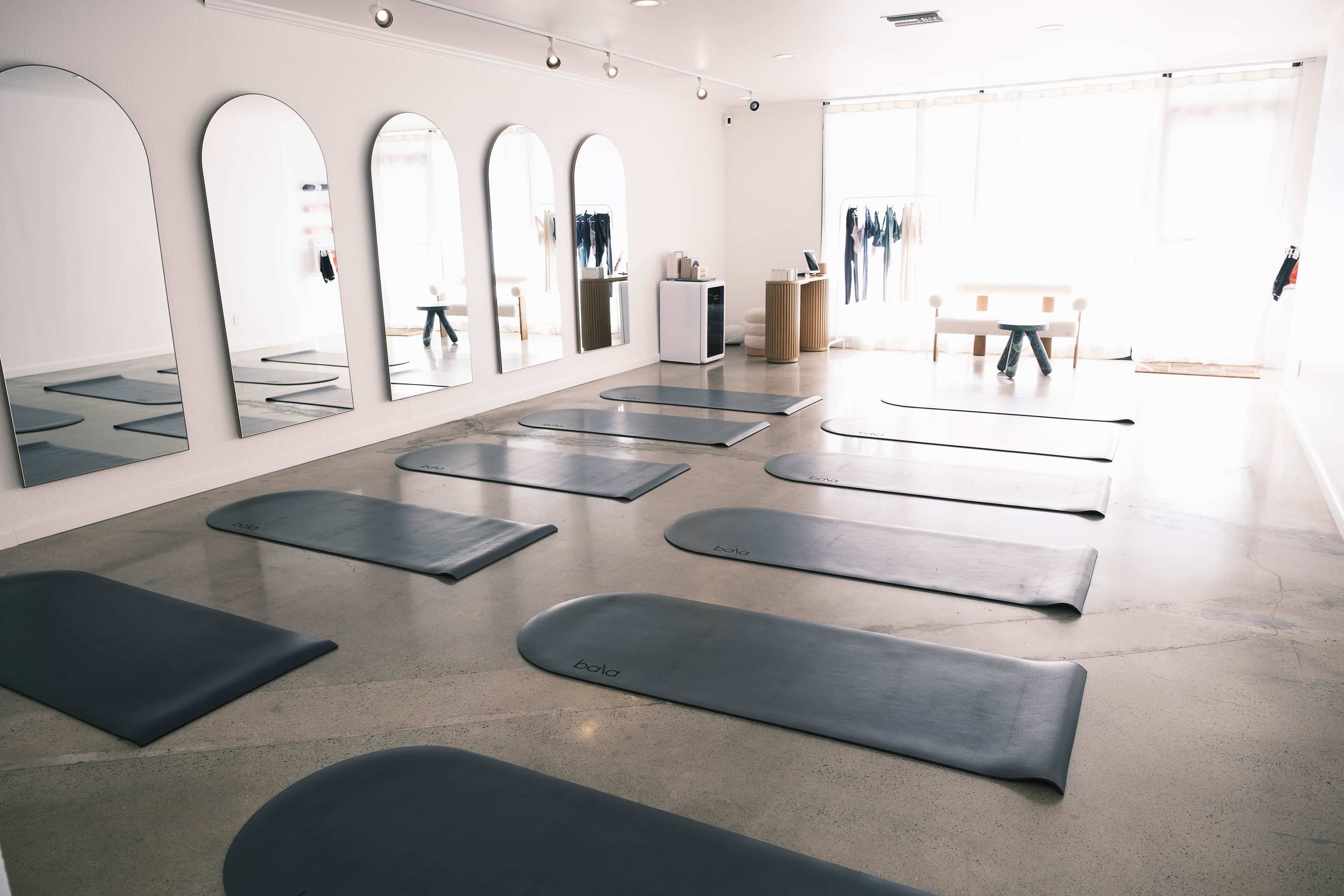 A spacious yoga studio features several yoga mats arranged in rows, mirrored walls, and natural light streaming in through large windows.