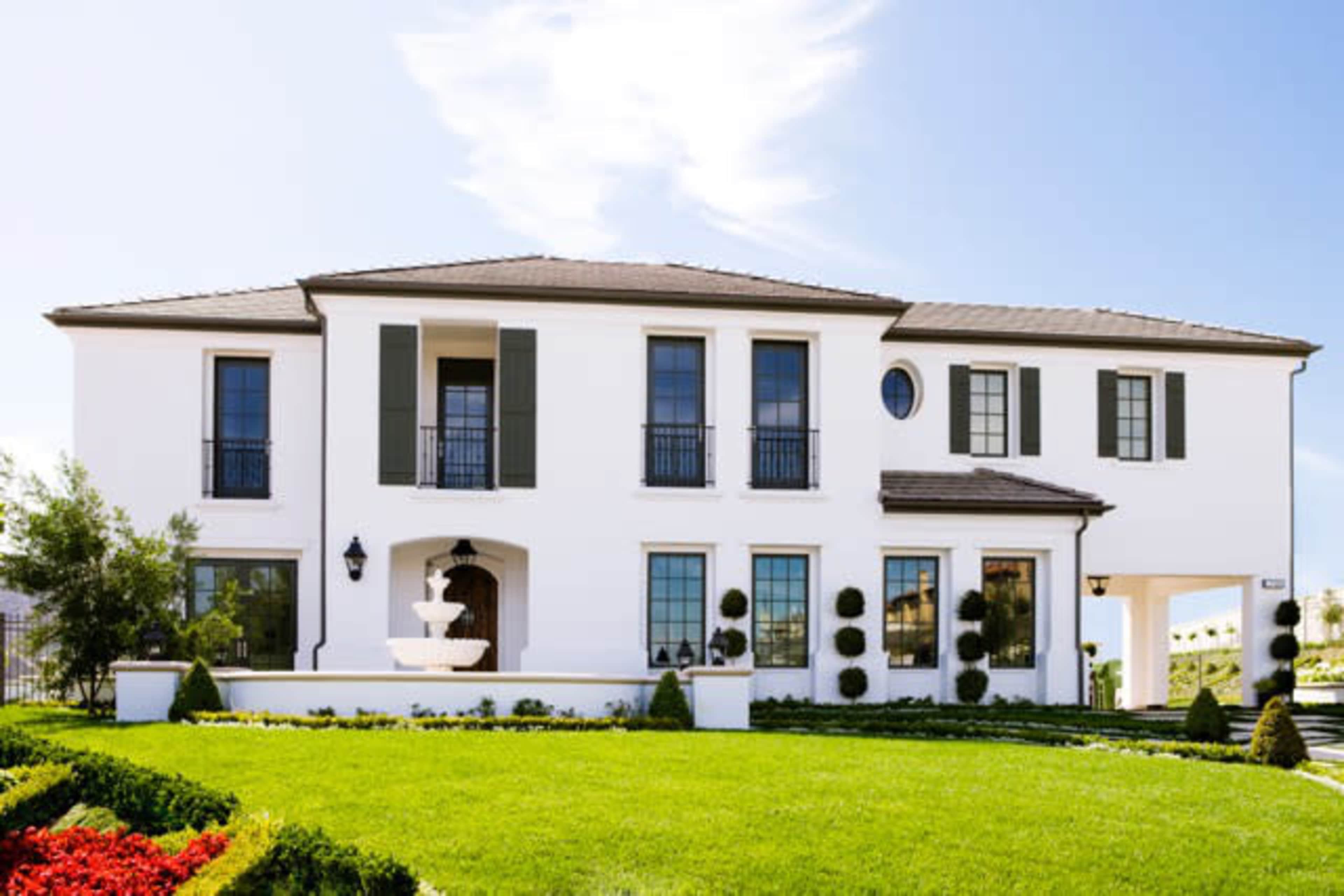 A modern two-story white house with green shutters, a fountain in the front, and neatly landscaped gardens.