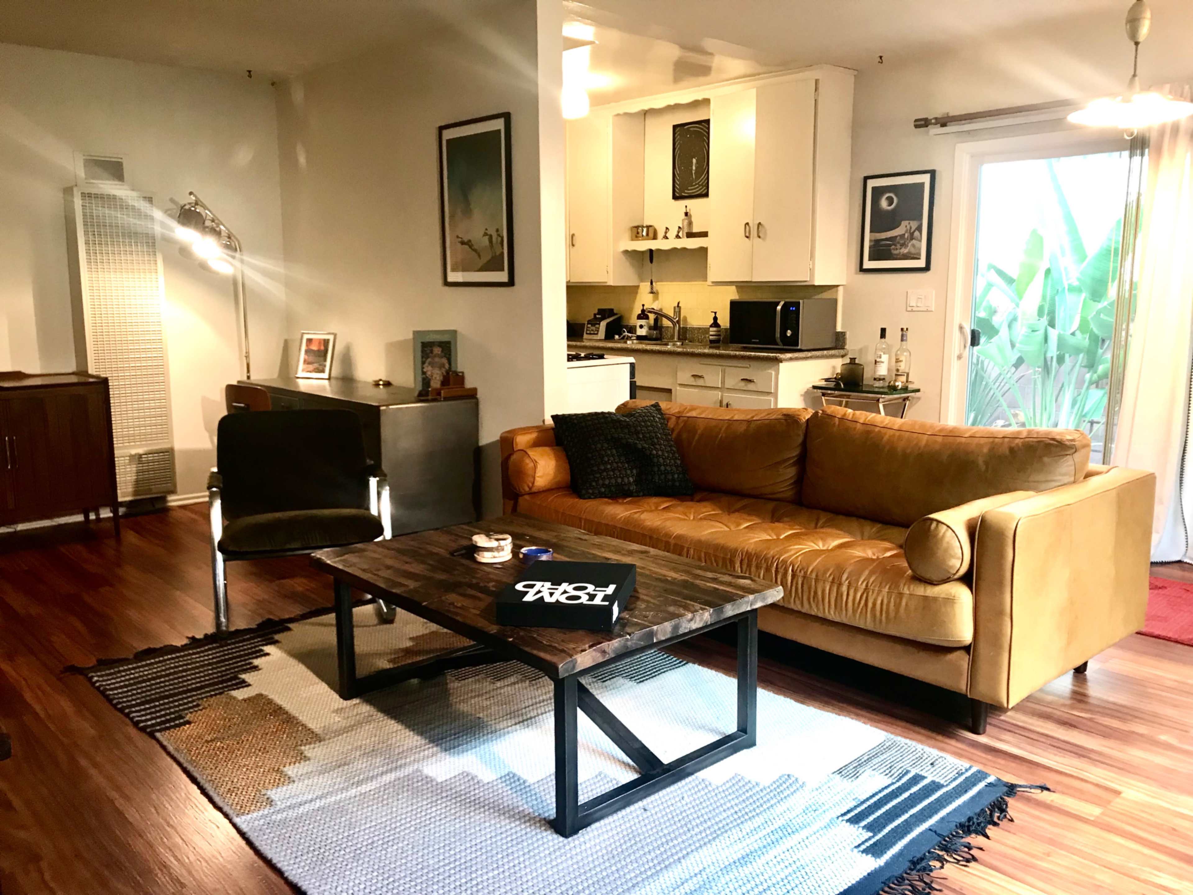 The image shows a cozy living room featuring a brown leather sofa, a wooden coffee table, and a modern kitchen area in the background.