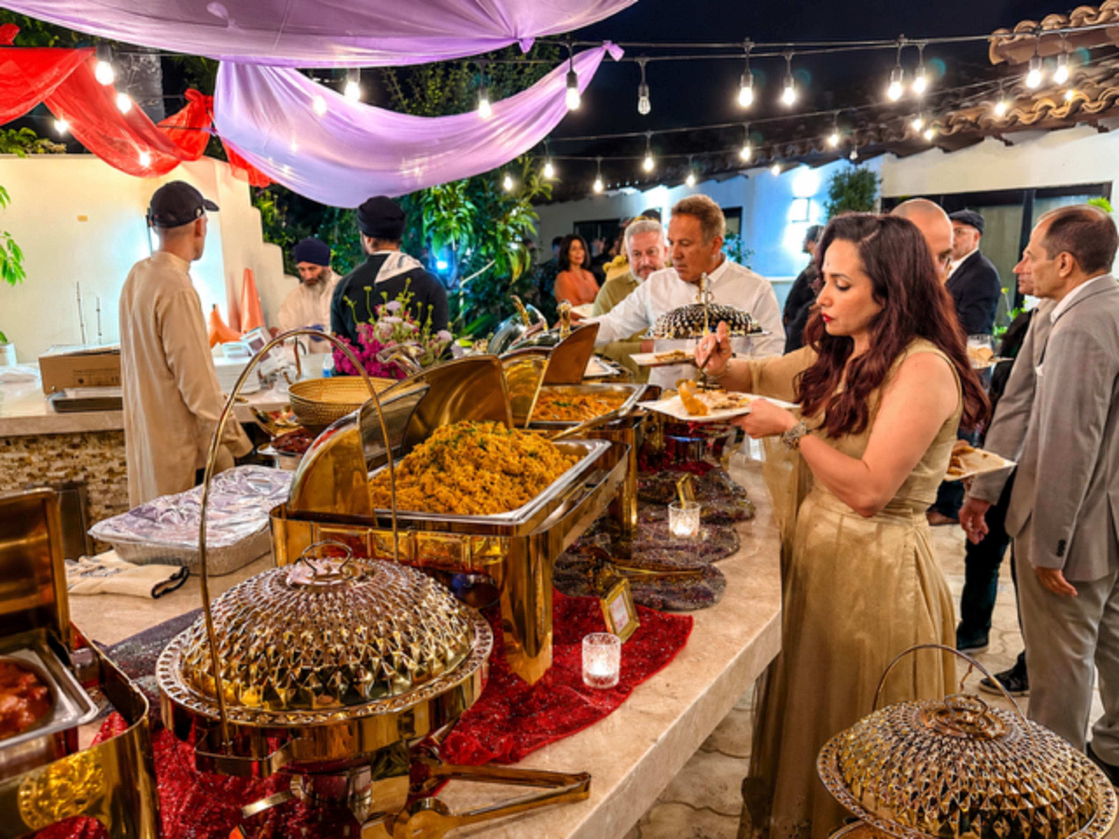 A buffet is set up outdoors with guests serving themselves food under string lights and draped fabric.
