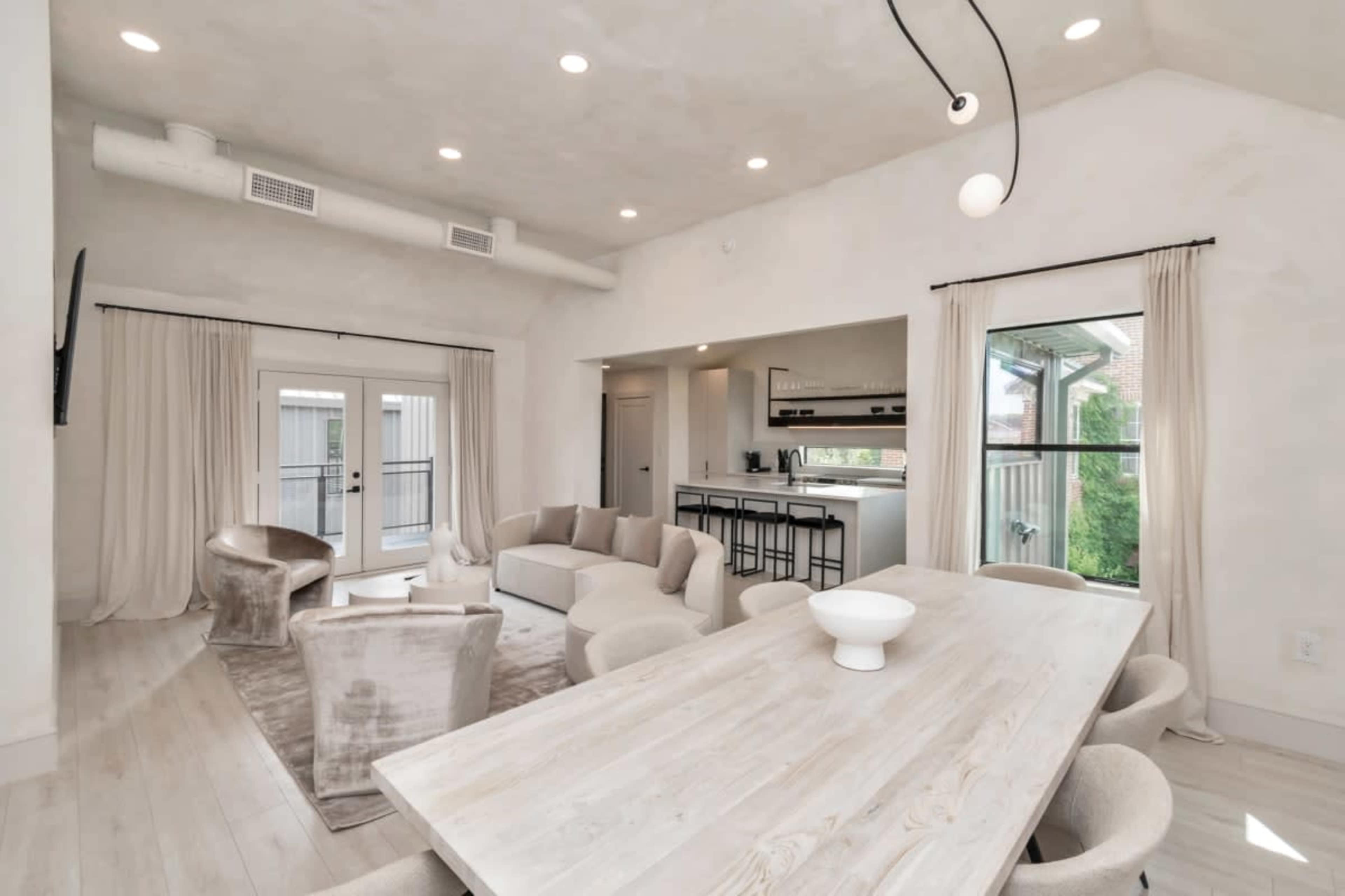 The image shows a modern, open-concept living space with a light-colored interior, featuring a dining table, comfortable seating, and large windows leading to a balcony.