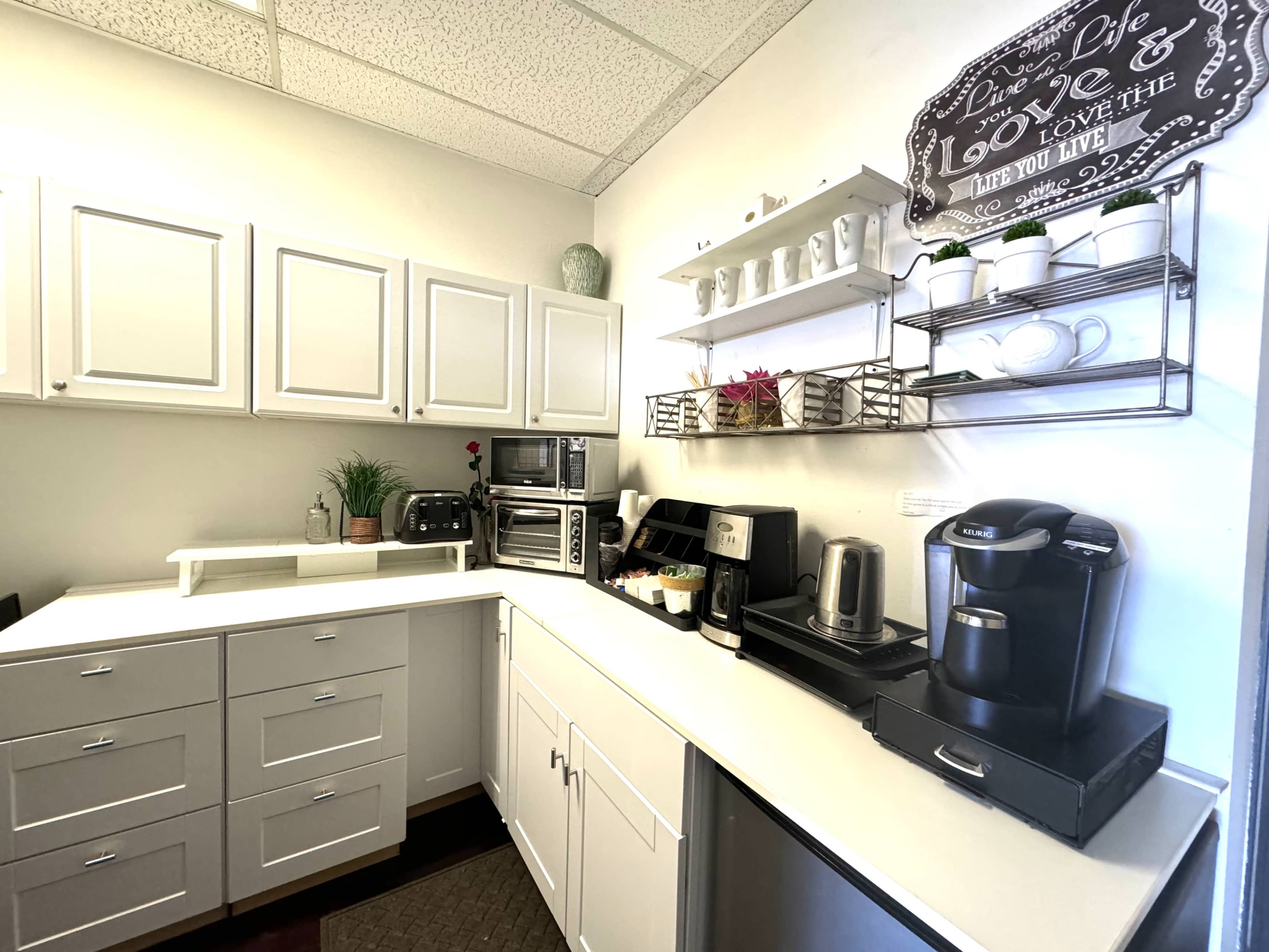 A compact kitchen features white cabinets, a countertop with various appliances, a wire shelf displaying cups and plants, and an organized arrangement of kitchen items.