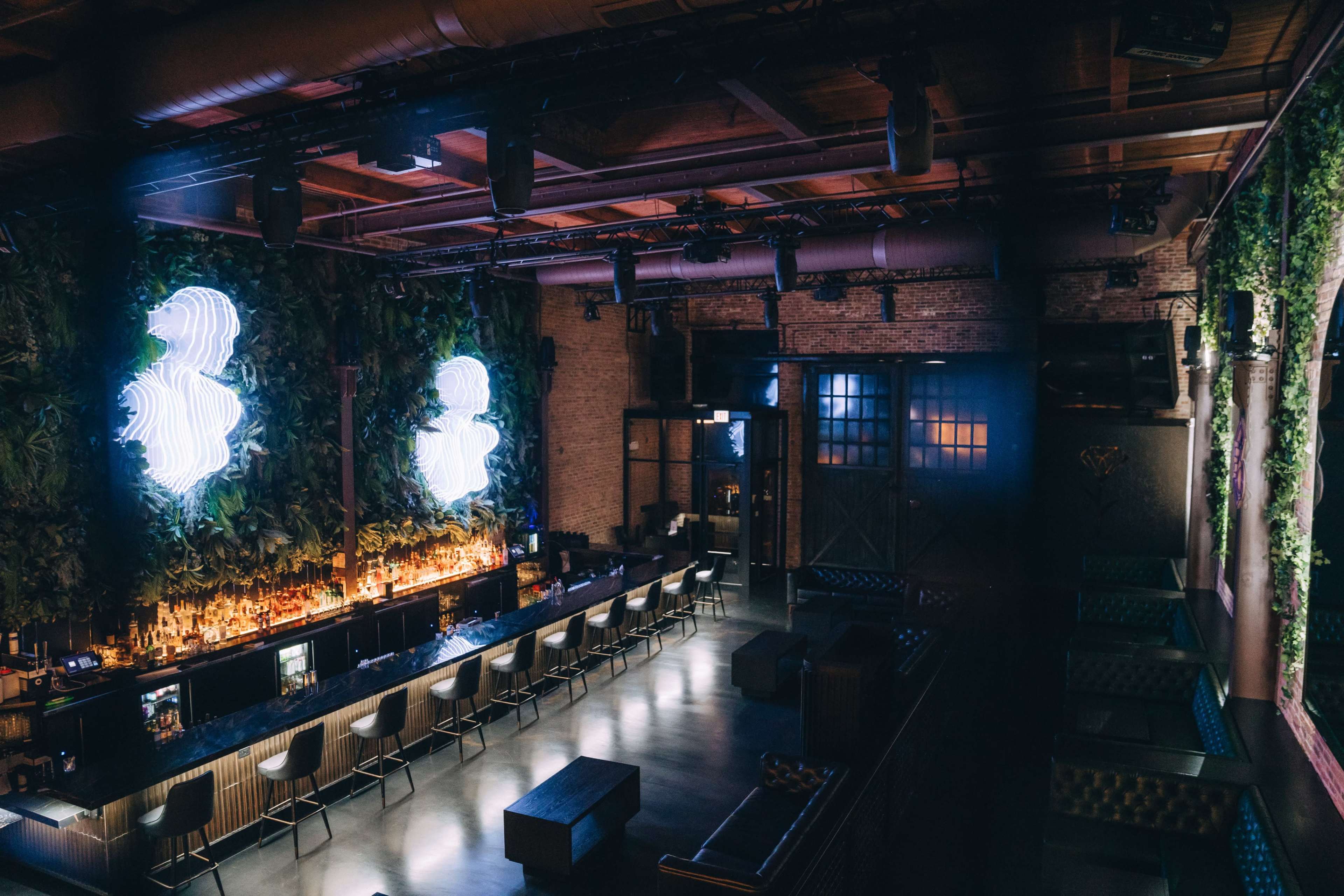 The image shows an upscale bar interior with a lush green wall, illuminated neon signs, and a long bar counter with stools.