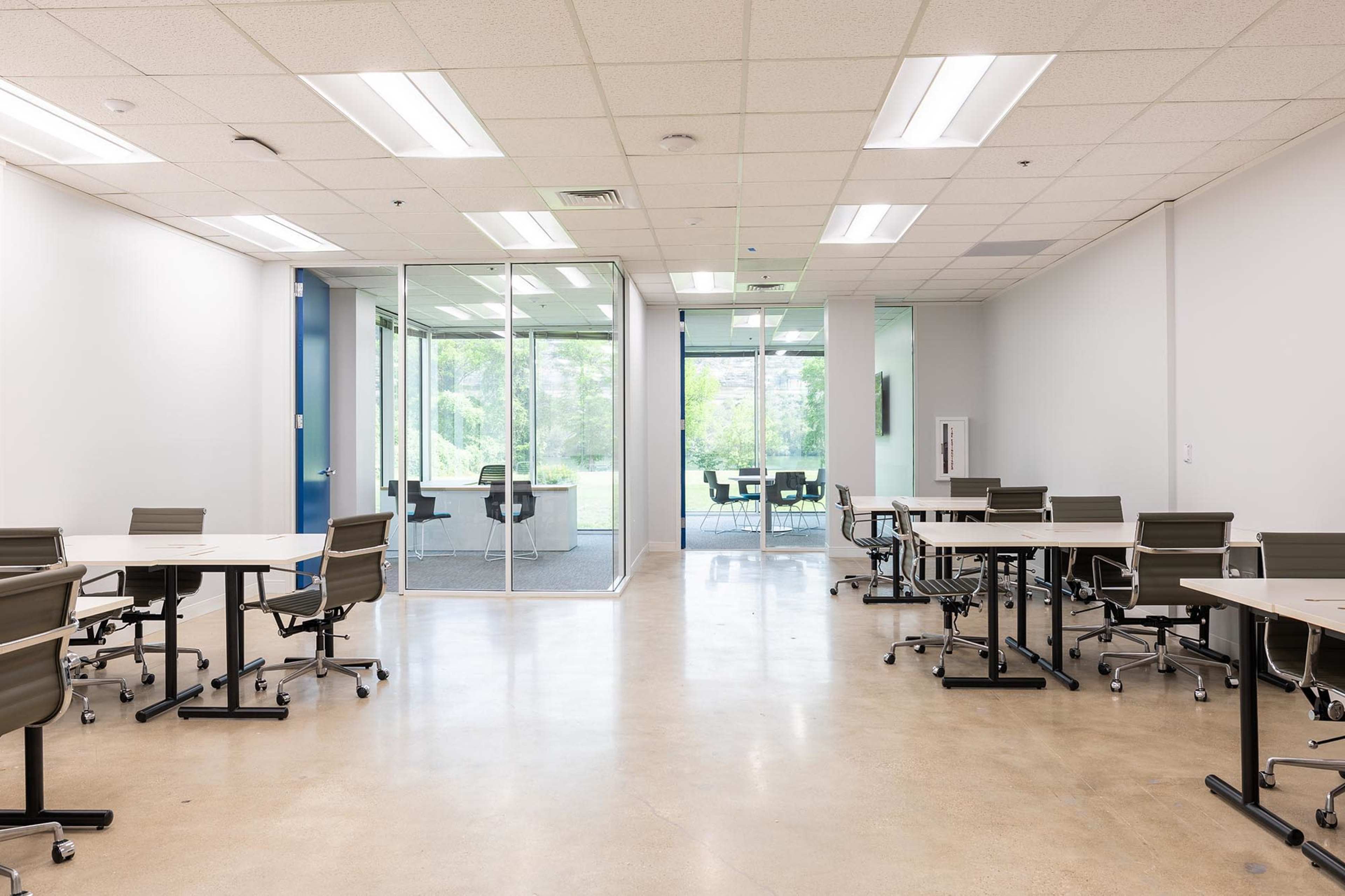 The image shows a modern office space with several empty tables and chairs arranged in a room that opens to a glass-enclosed meeting area.