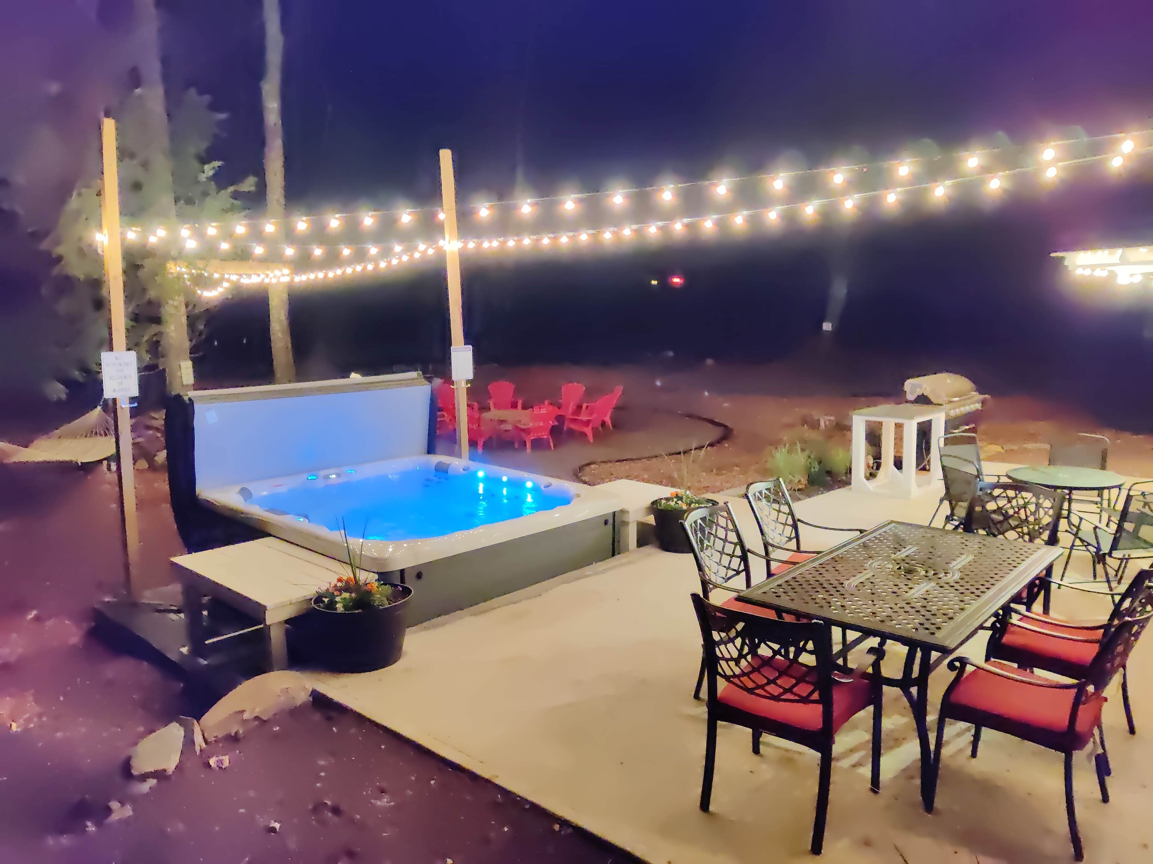 The image shows a backyard area at night featuring a hot tub, a dining table with chairs, and string lights overhead.
