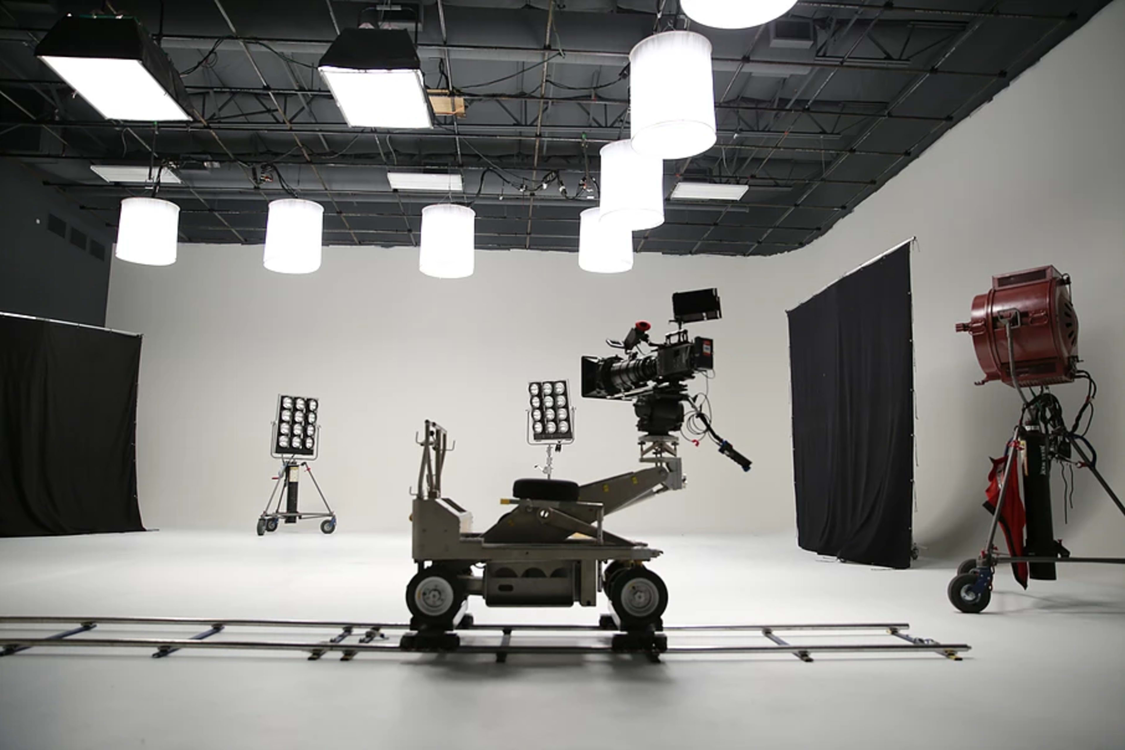 A film studio set features camera equipment on a dolly track, mounted lights, and backdrop curtains.