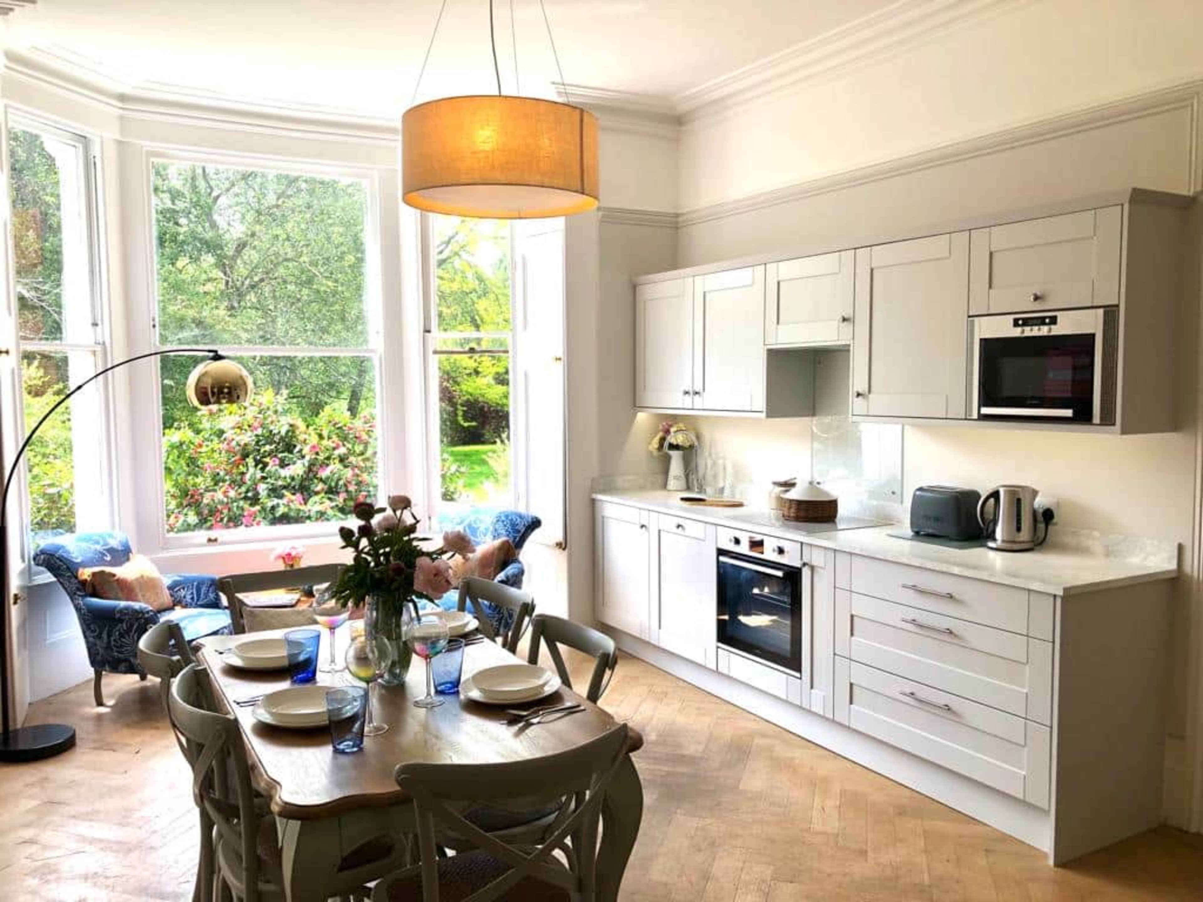 A modern kitchen with white cabinetry and a wooden dining table is set beside large windows overlooking a green garden.