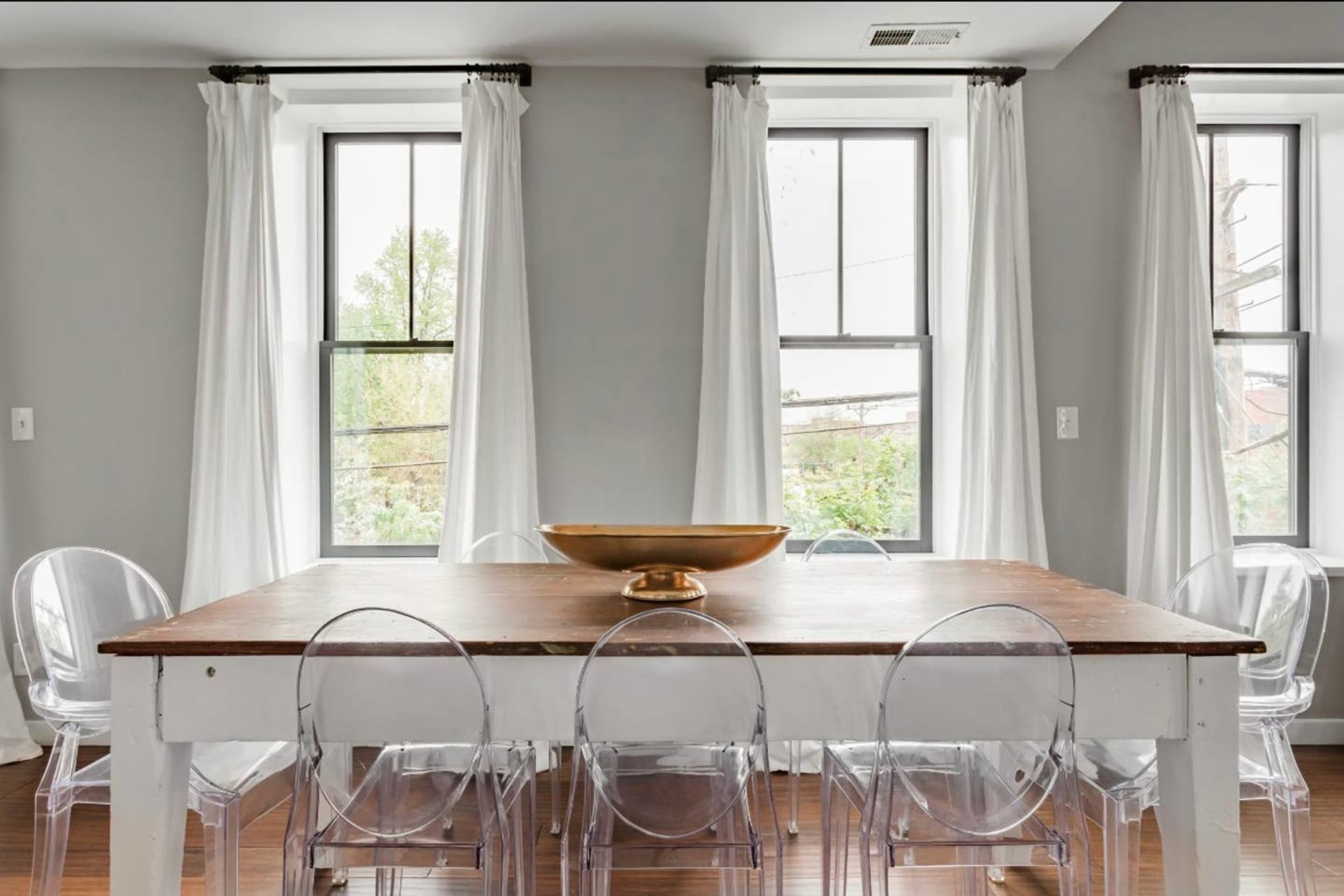 A dining table with a bowl at the center is surrounded by several clear chairs, positioned near windows in a well-lit room.