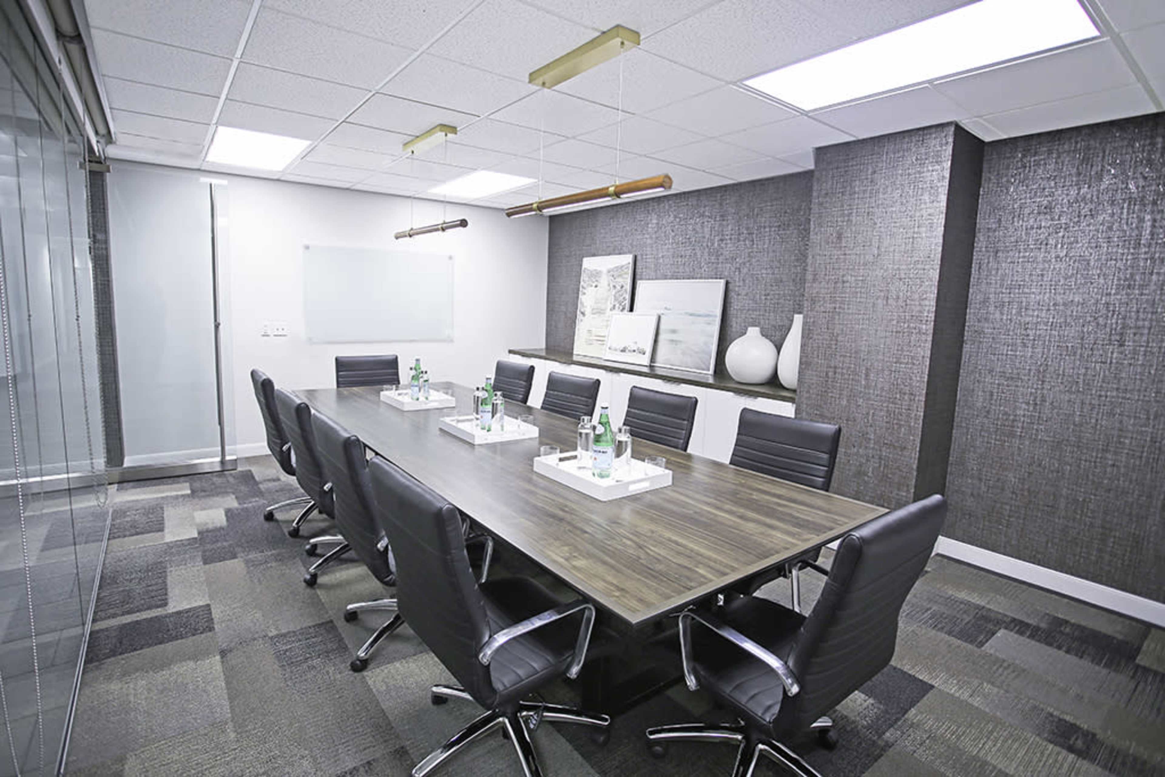 The image shows a modern conference room with a long wooden table surrounded by black leather chairs, featuring two water bottles on trays at each seating space.