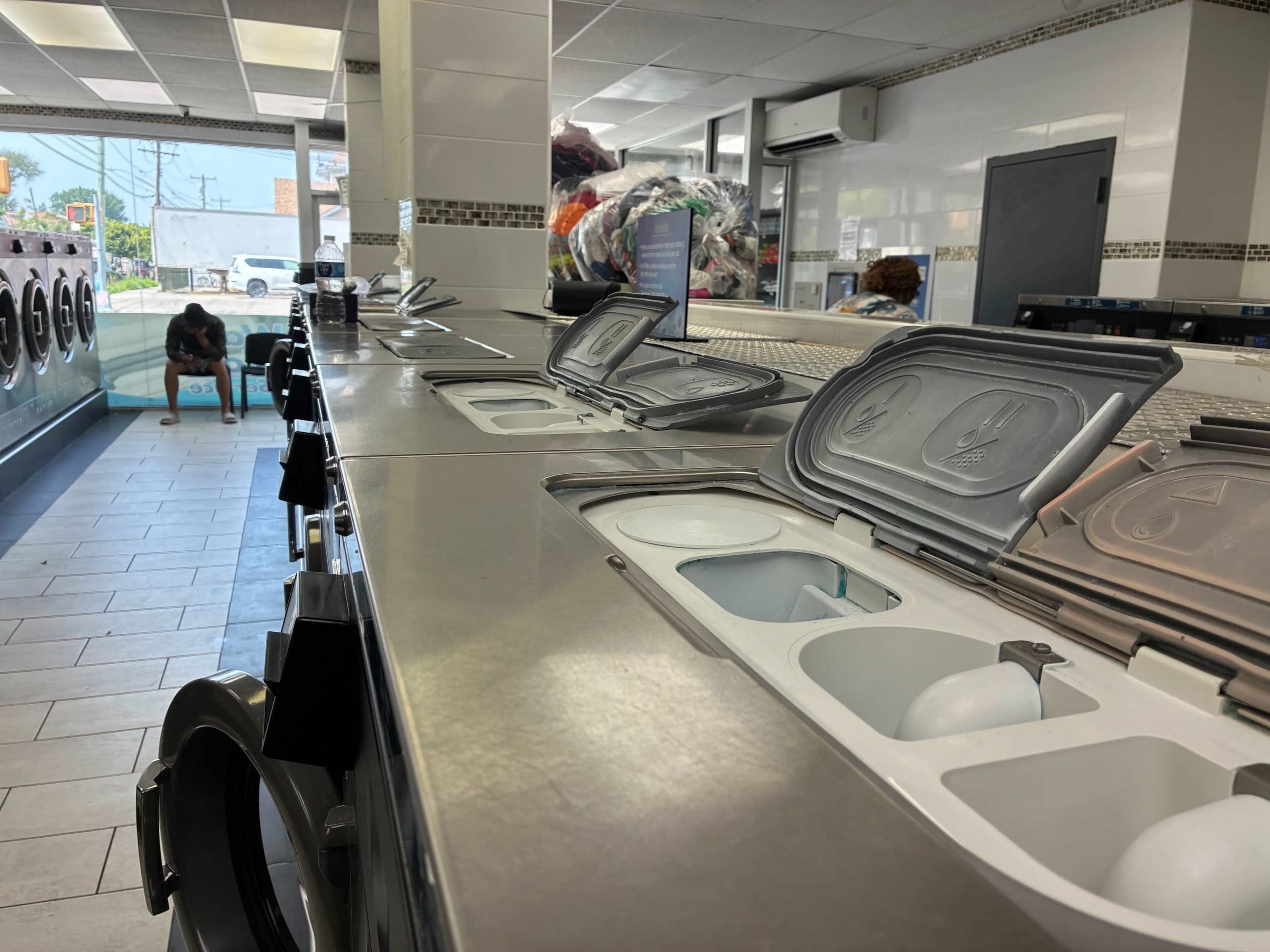 The image shows the interior of a laundromat with metal washing machines lined up and a person sitting on a bench in the background.