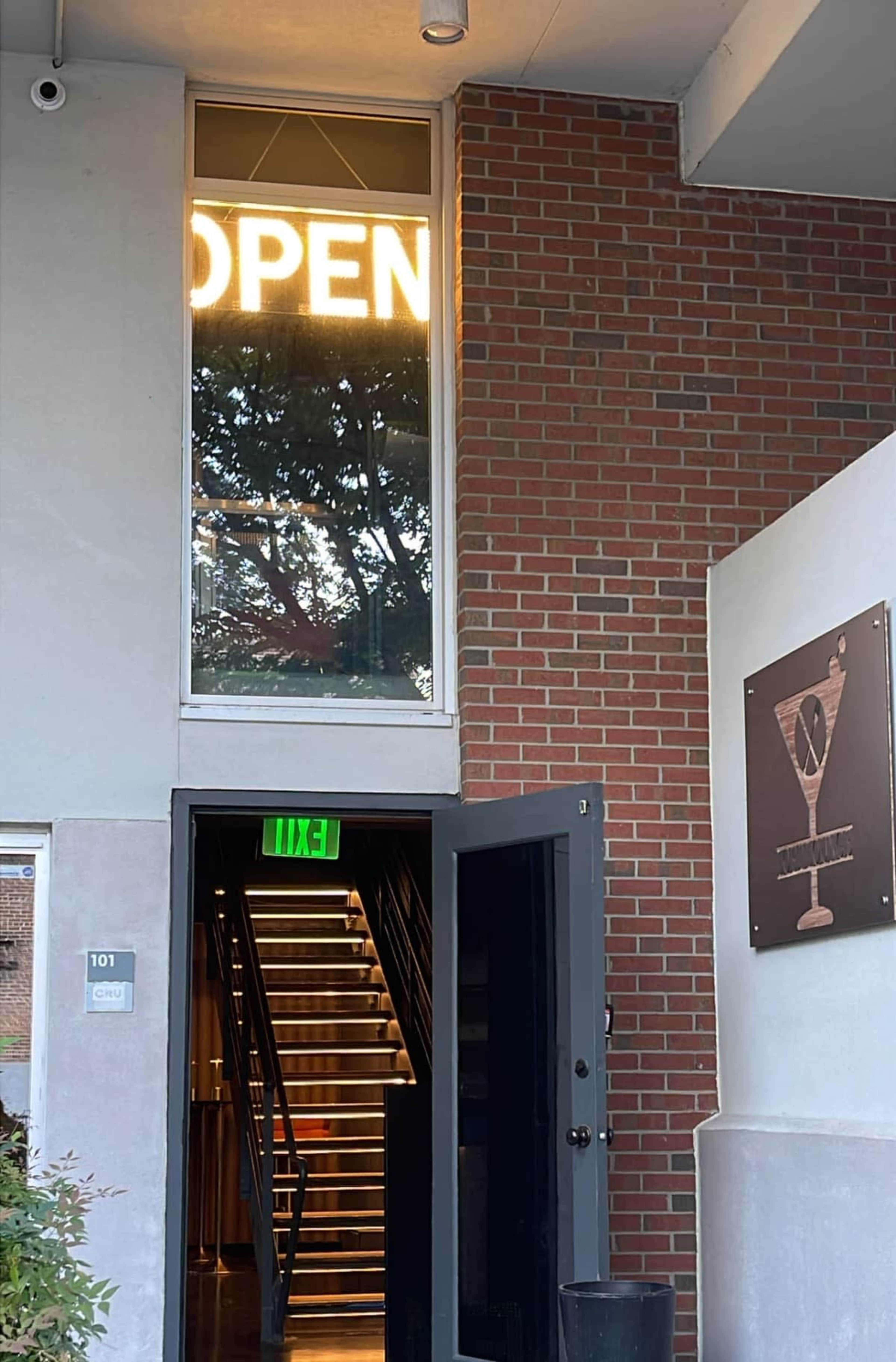A glass door stands open, revealing illuminated "OPEN" signage above a staircase leading upwards inside a brick building.