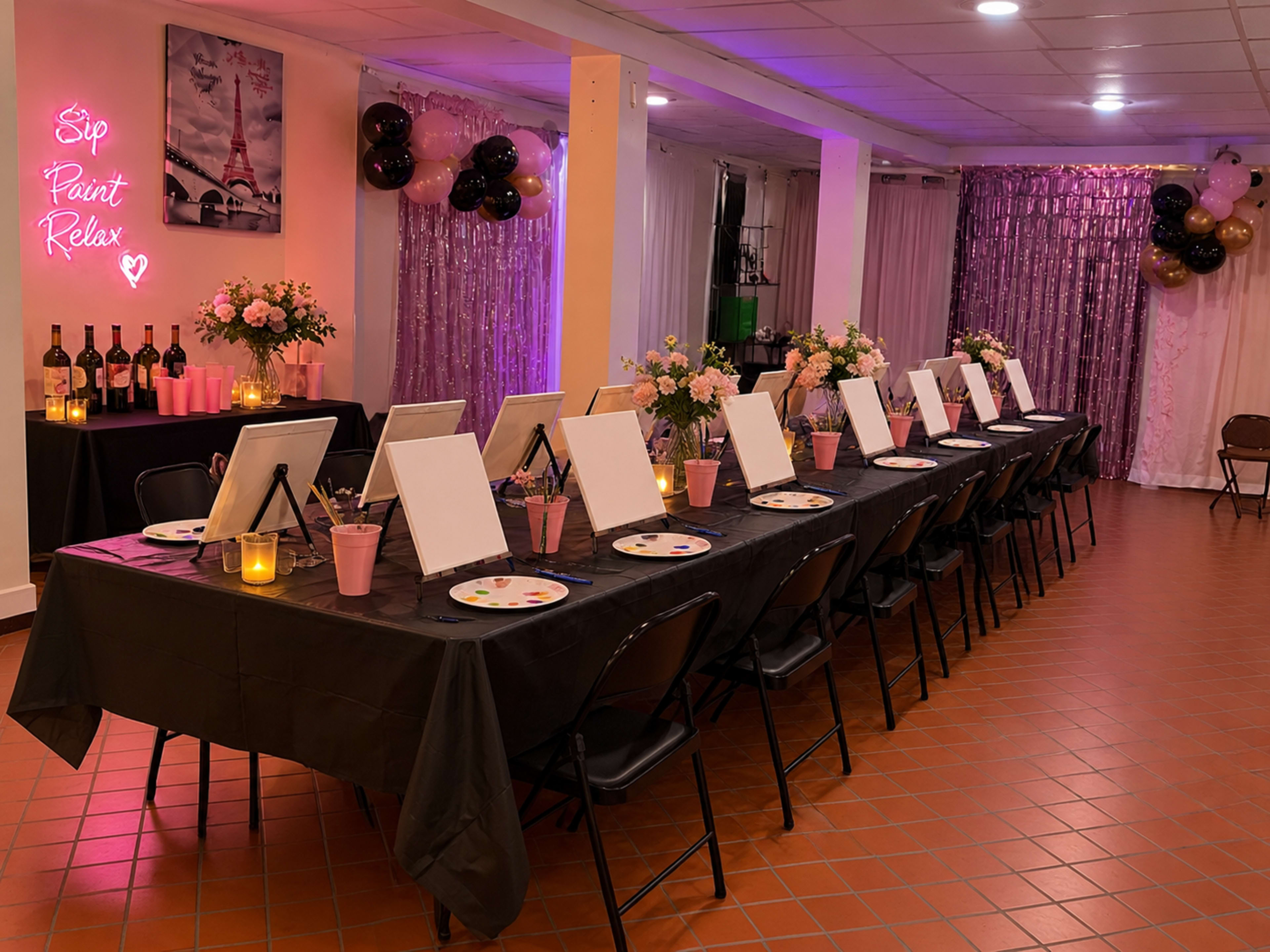 The image shows a decorated event space with a long table set for a painting party, featuring blank canvases, paint supplies, wine bottles, and floral centerpieces under soft purple lighting.