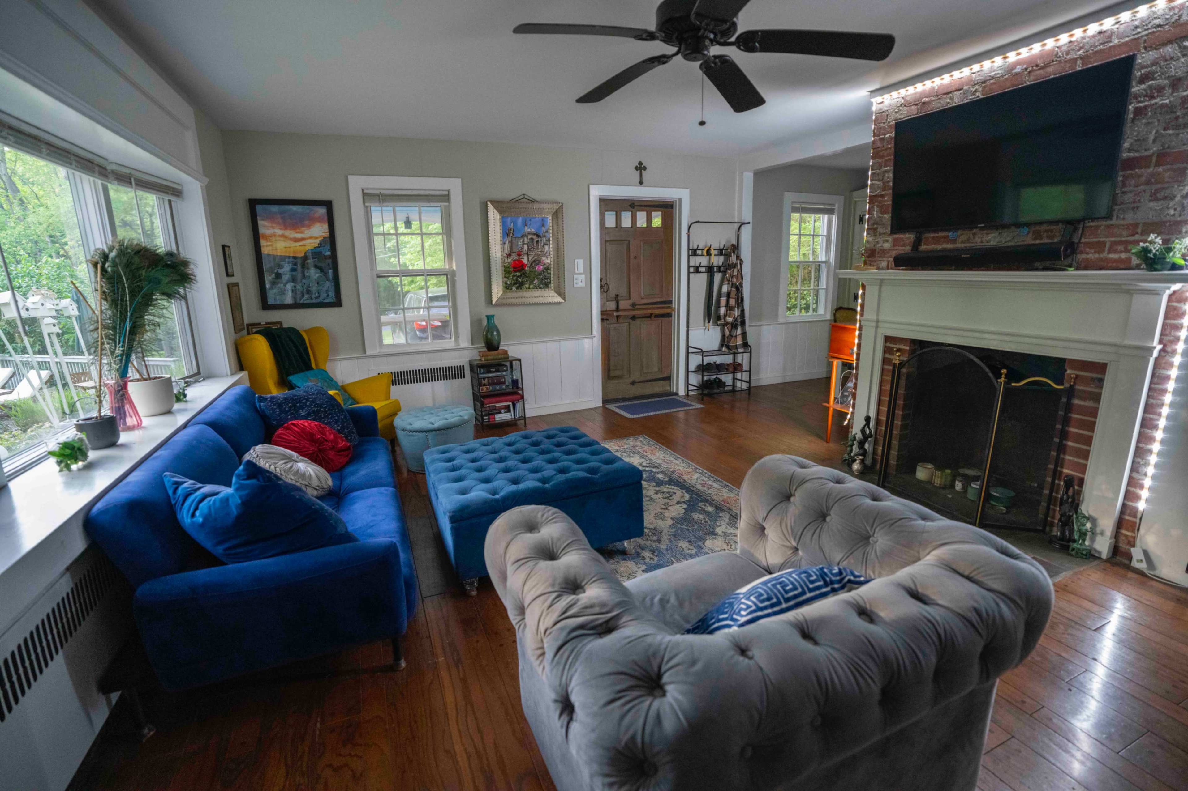 The image shows a living room with blue sofas, a tufted ottoman, and a fireplace, surrounded by large windows and various decorative elements.