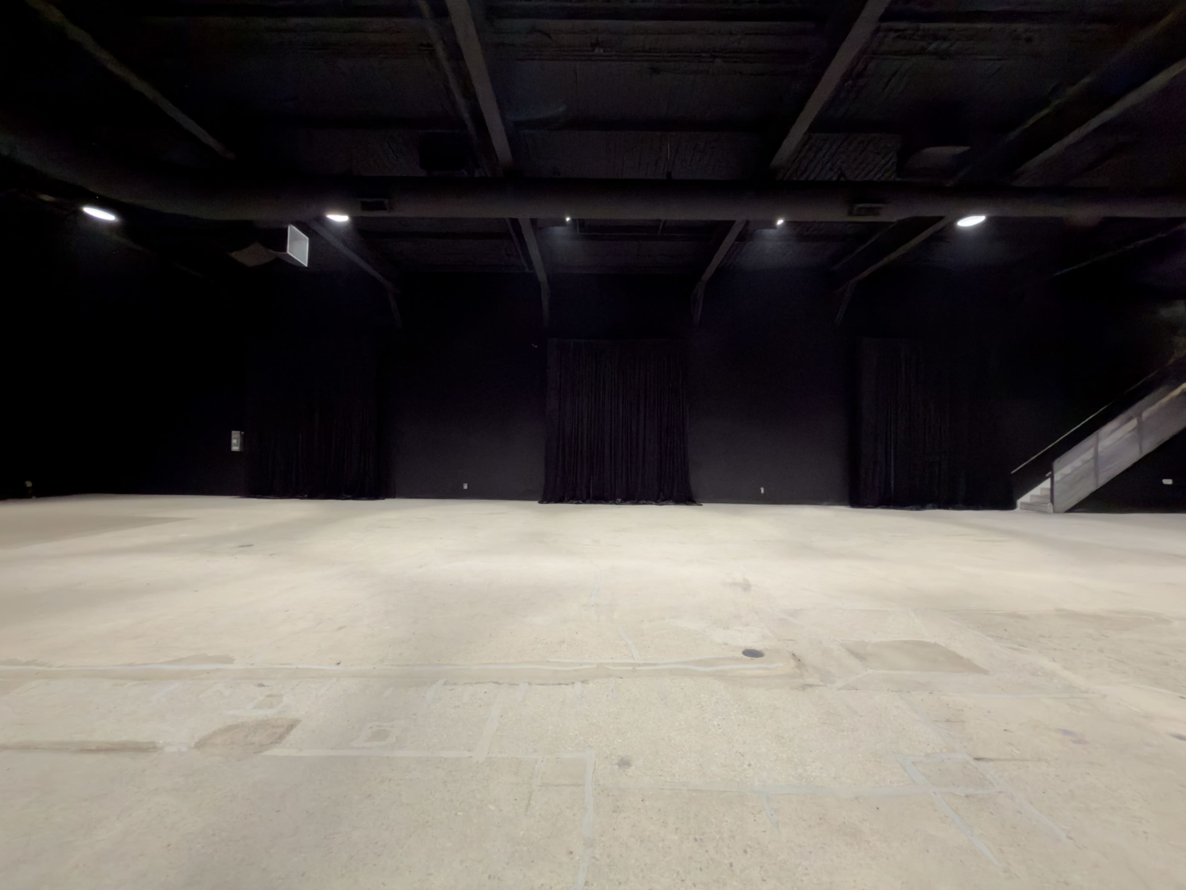 The image shows an empty dark room with black walls and a concrete floor, featuring two black curtains and a staircase in the corner.