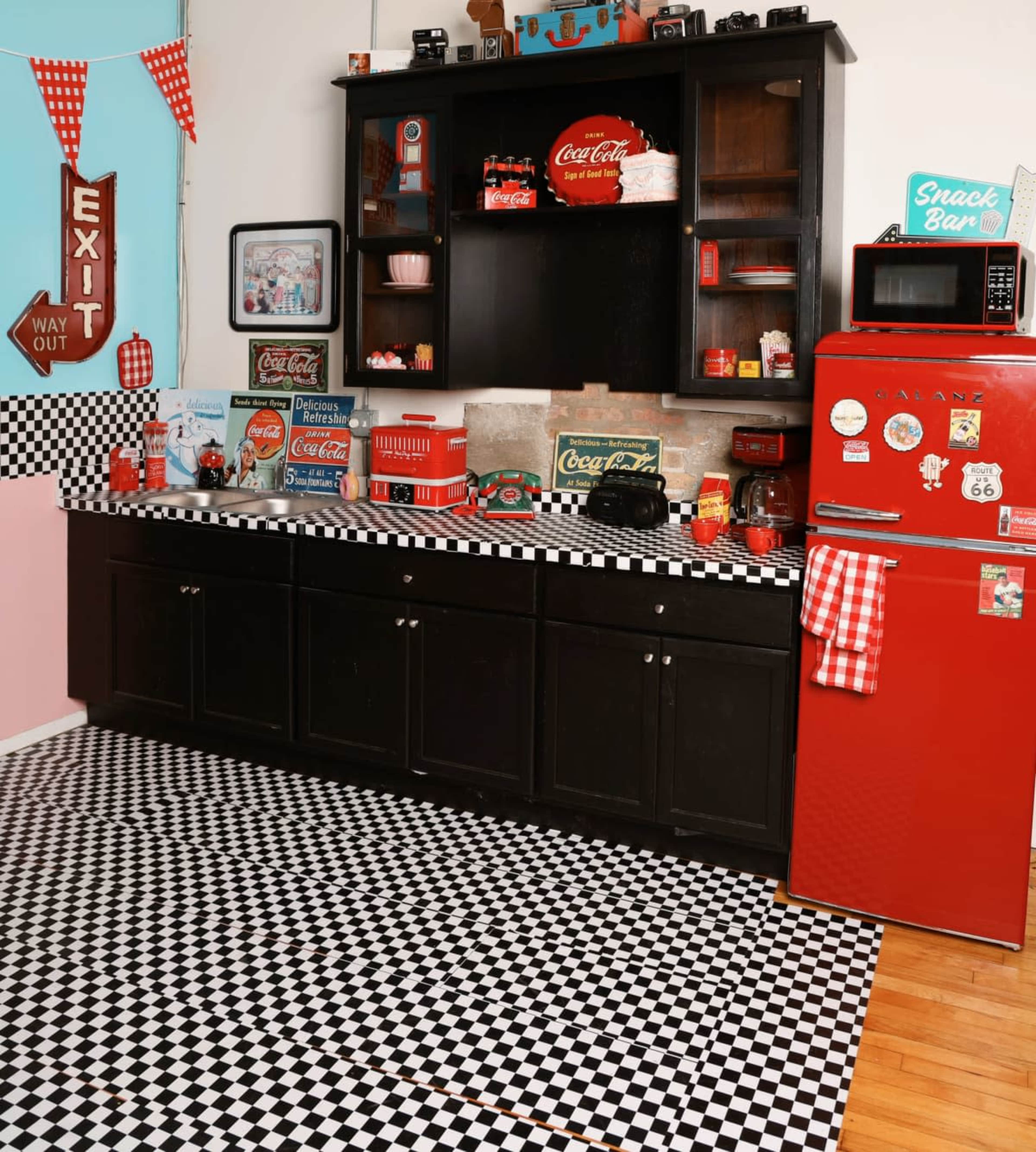 The image shows a vintage-themed kitchen with a checkered floor, black cabinetry, a red refrigerator, and various Coca-Cola memorabilia on display.