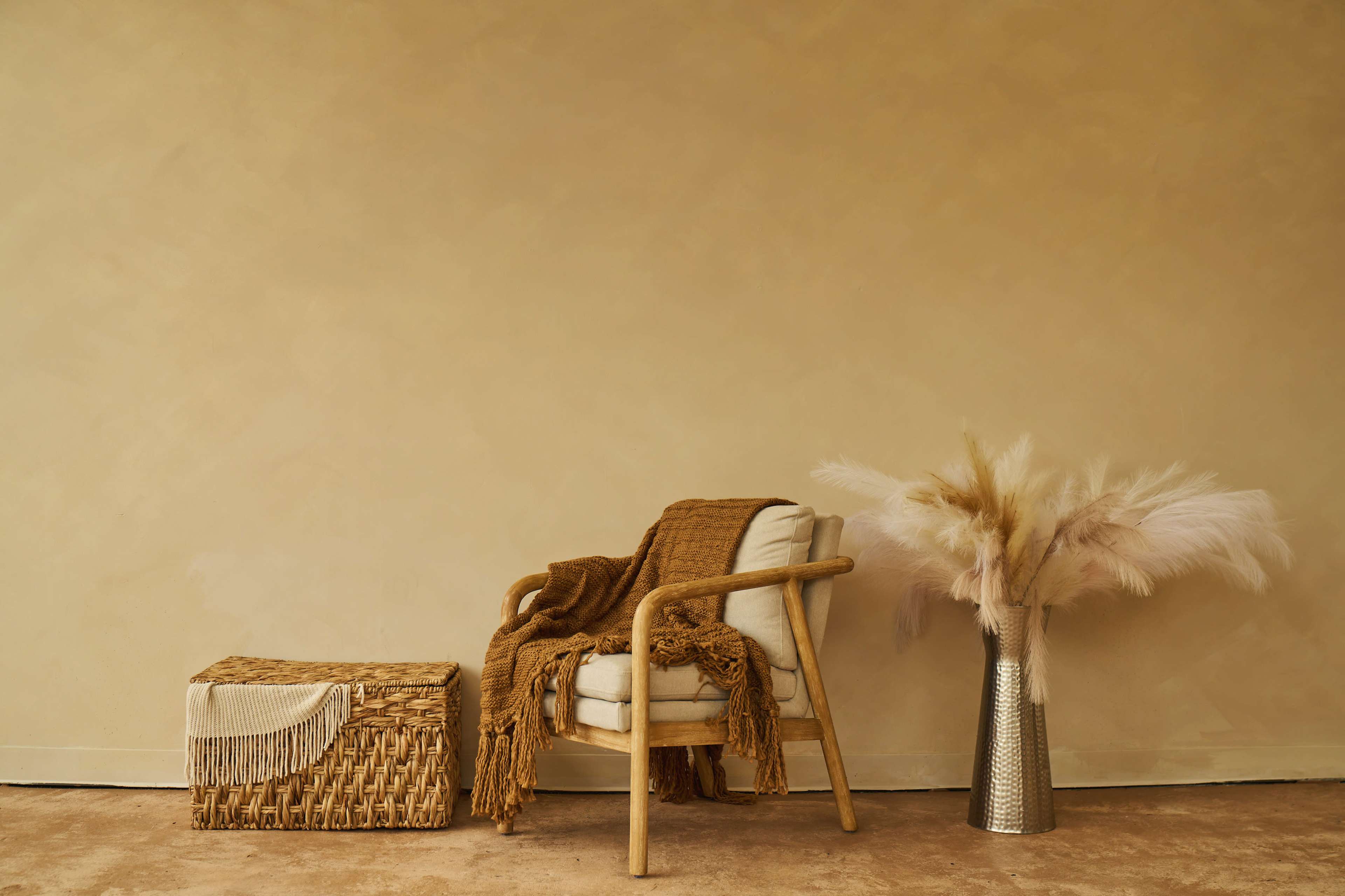A wooden chair with a tan throw and a woven storage box sit beside a vase of dried pampas grass against a plain beige wall.