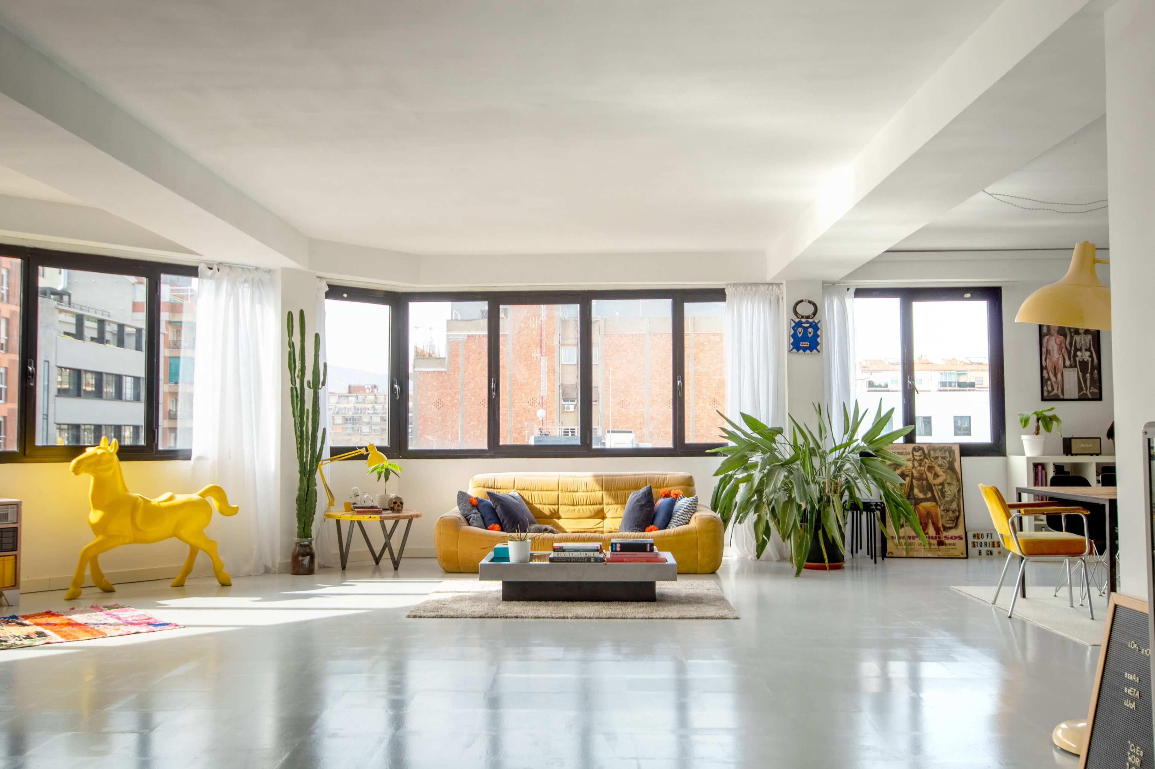 A bright, modern living room with large windows, a yellow sofa, a coffee table, plants, and a yellow decorative horse.