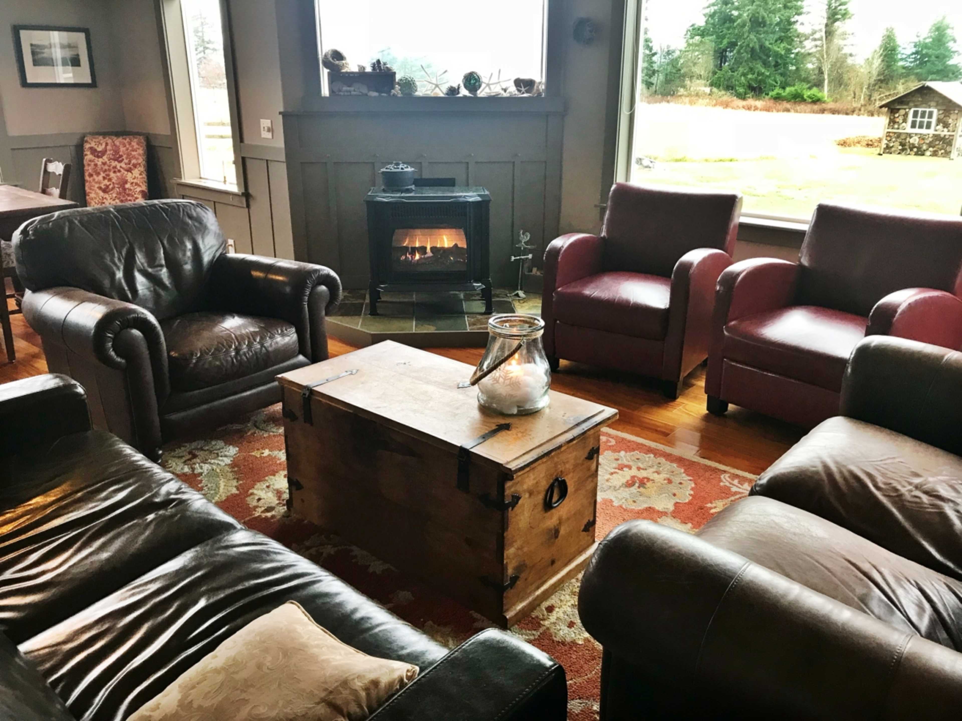 The scene depicts a cozy living room with leather sofas, a wooden coffee table, and a fireplace, surrounded by large windows overlooking a green landscape.