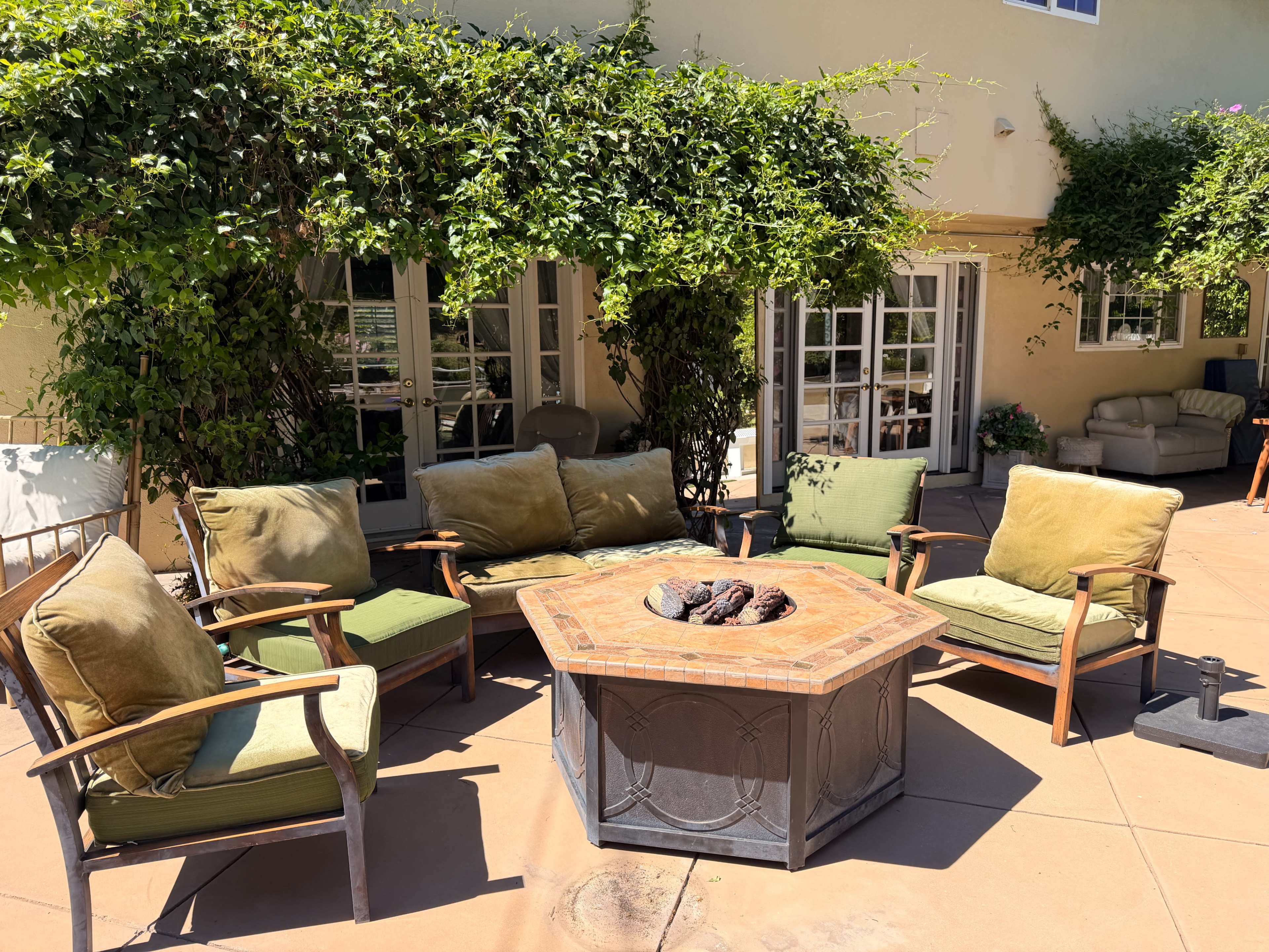 A circular stone fire pit surrounded by green and brown cushioned chairs on a patio with a backdrop of a light-colored building and leafy vines.