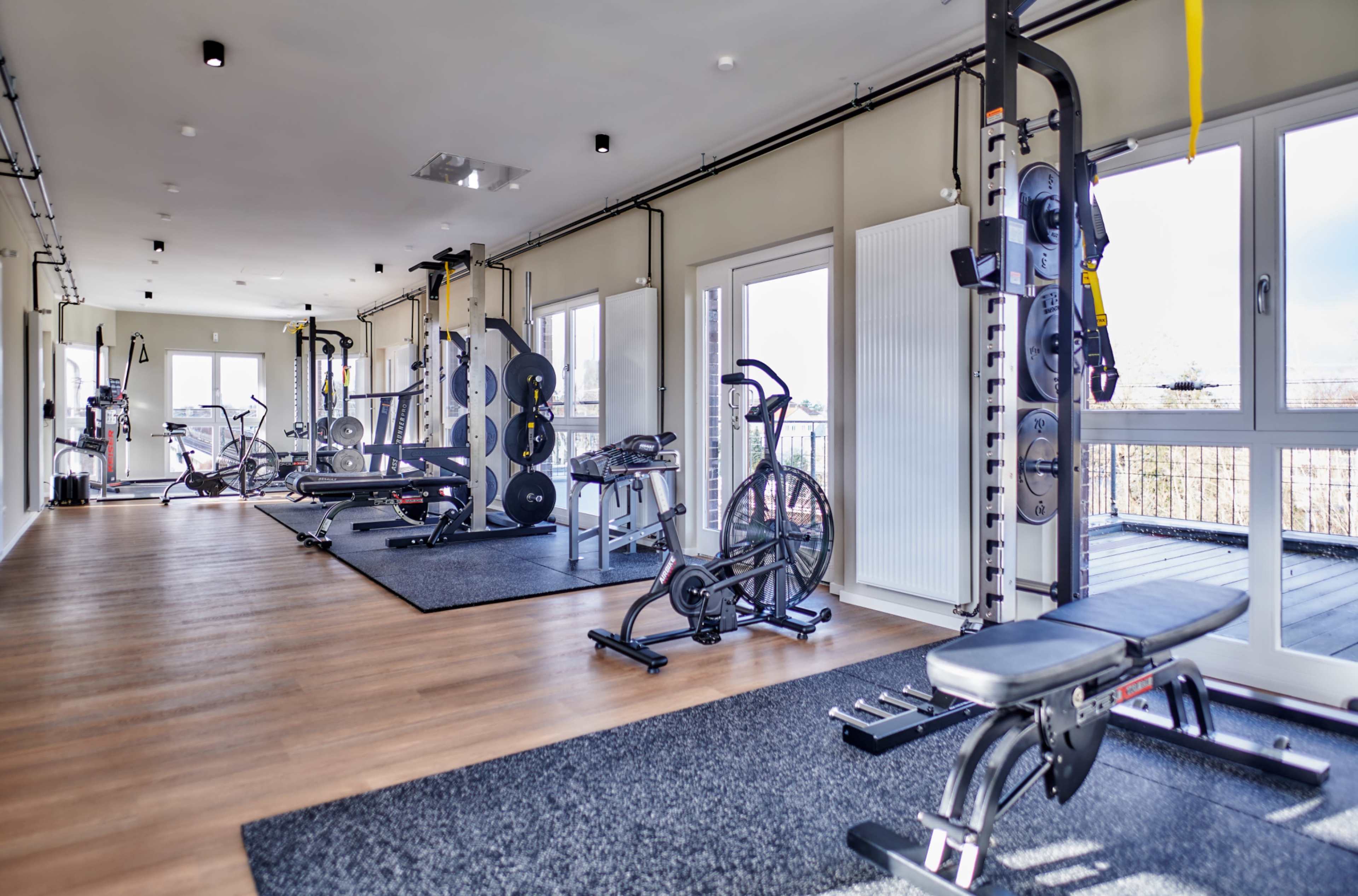 The image shows a well-equipped gym with various exercise machines and weights, featuring large windows that allow natural light.
