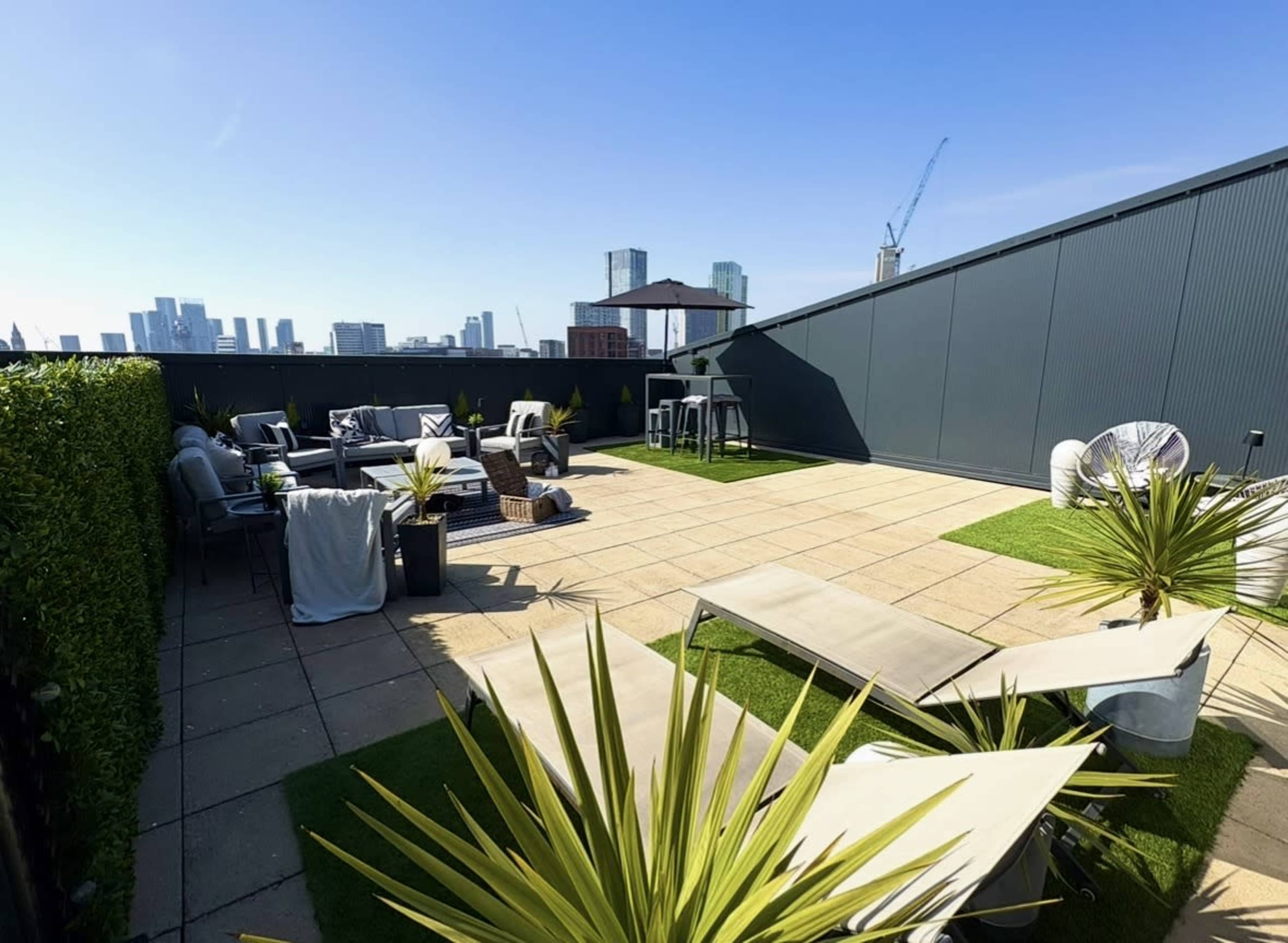 The image shows a rooftop terrace featuring lounge chairs, tables, and greenery, with a skyline view of city buildings in the background.