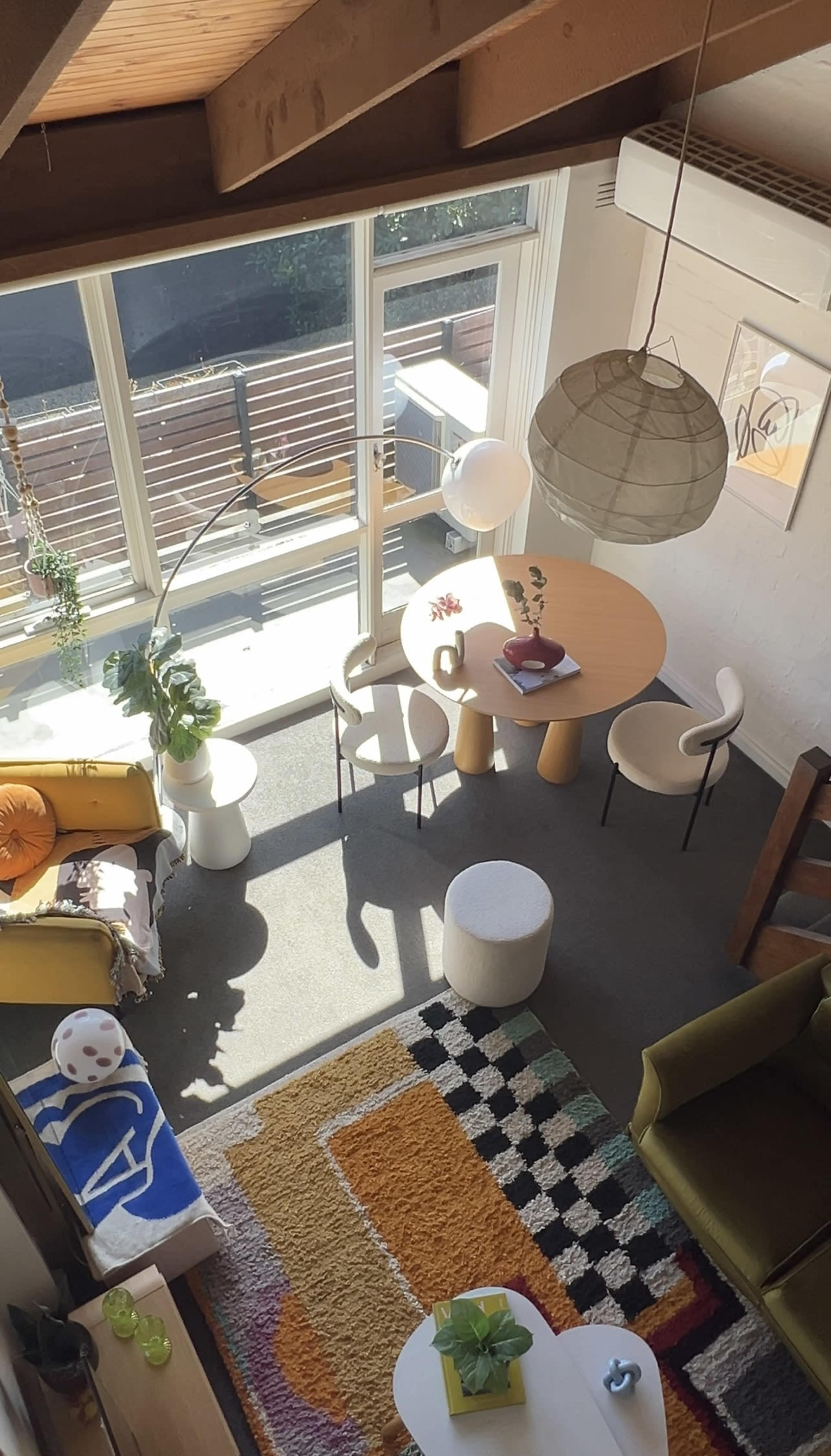 The image shows a brightly lit living space with a round dining table surrounded by chairs, a cozy seating area, and large windows offering a view of the outdoors.
