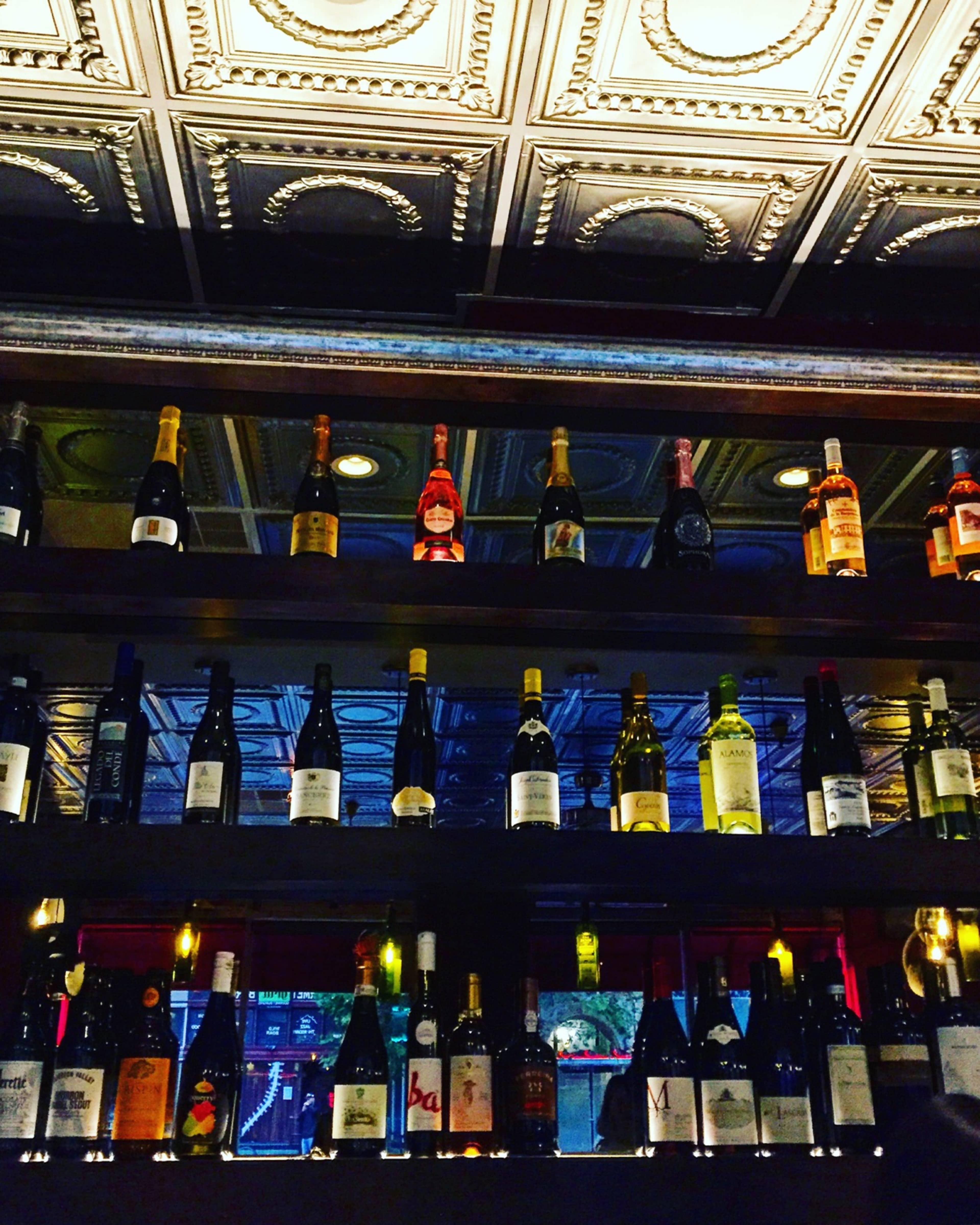 A row of wine bottles is neatly arranged on wooden shelves beneath a decorative tin ceiling.