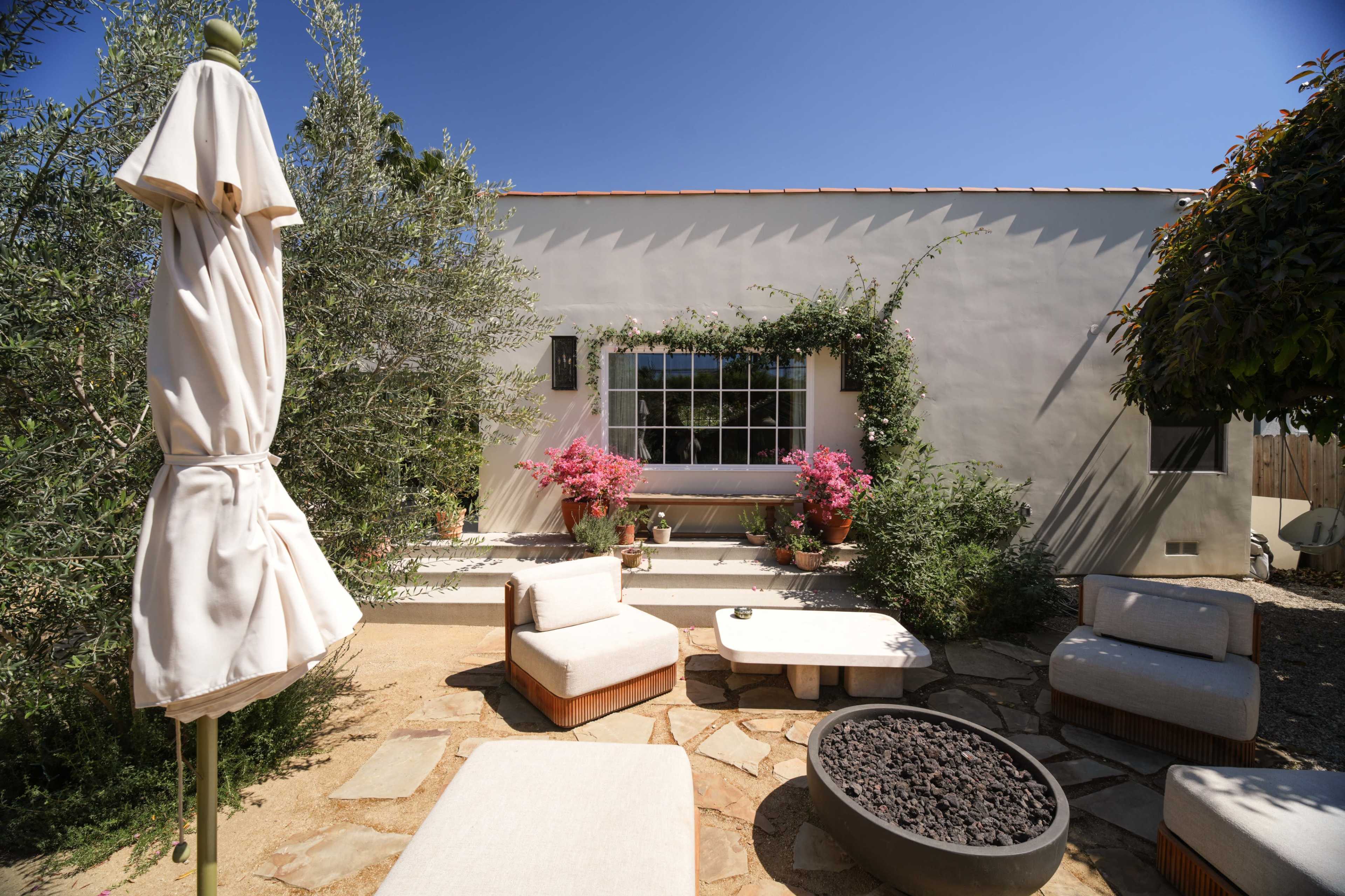 A cozy outdoor seating area features stone flooring, a fire pit, and decorative plants alongside a white-walled house.