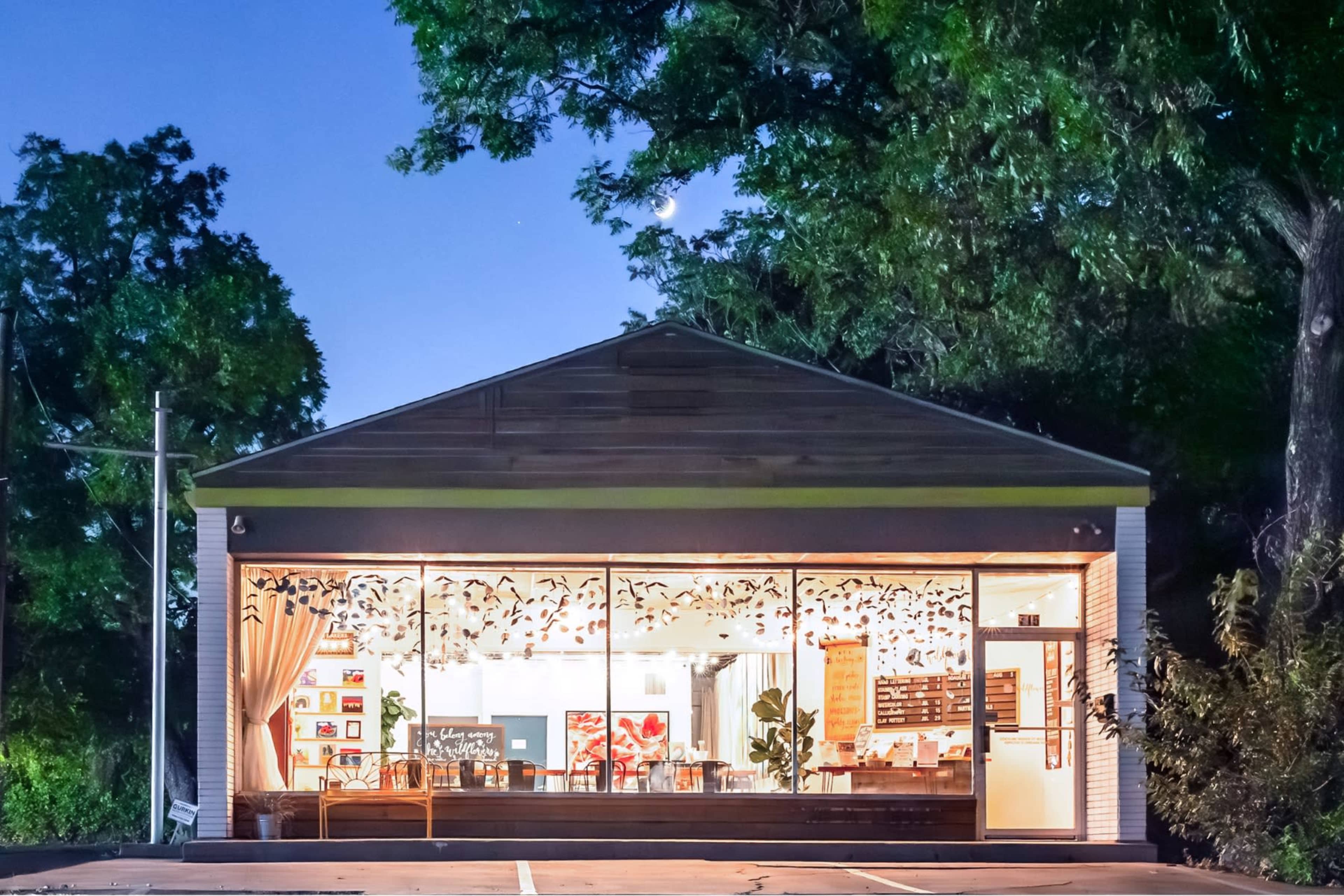 A brightly lit café with large front windows showcasing interior seating is set against a backdrop of trees at dusk.