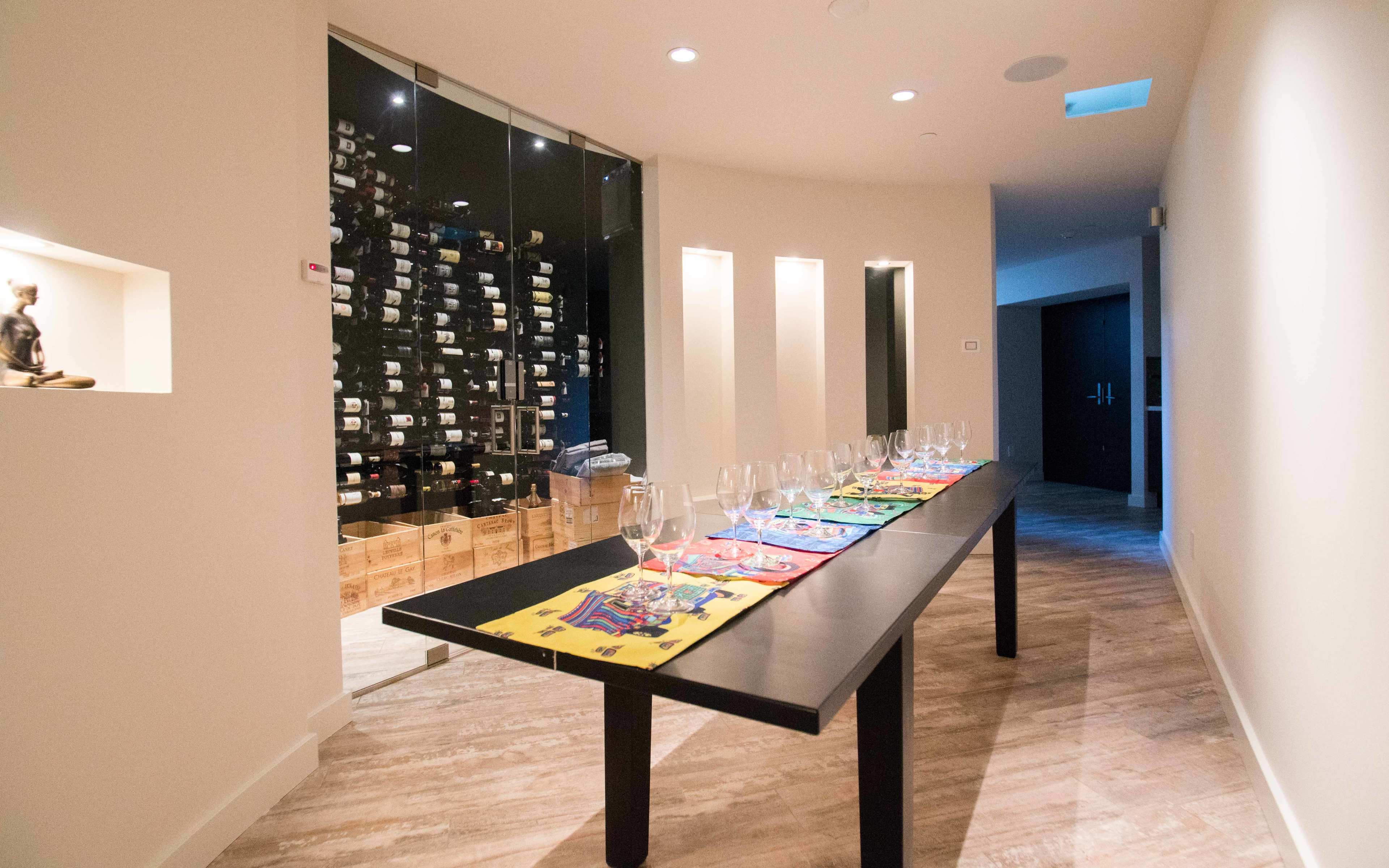 A long black table with colorful placemats is set up in a well-lit corridor next to a glass-enclosed wine storage area filled with bottles.
