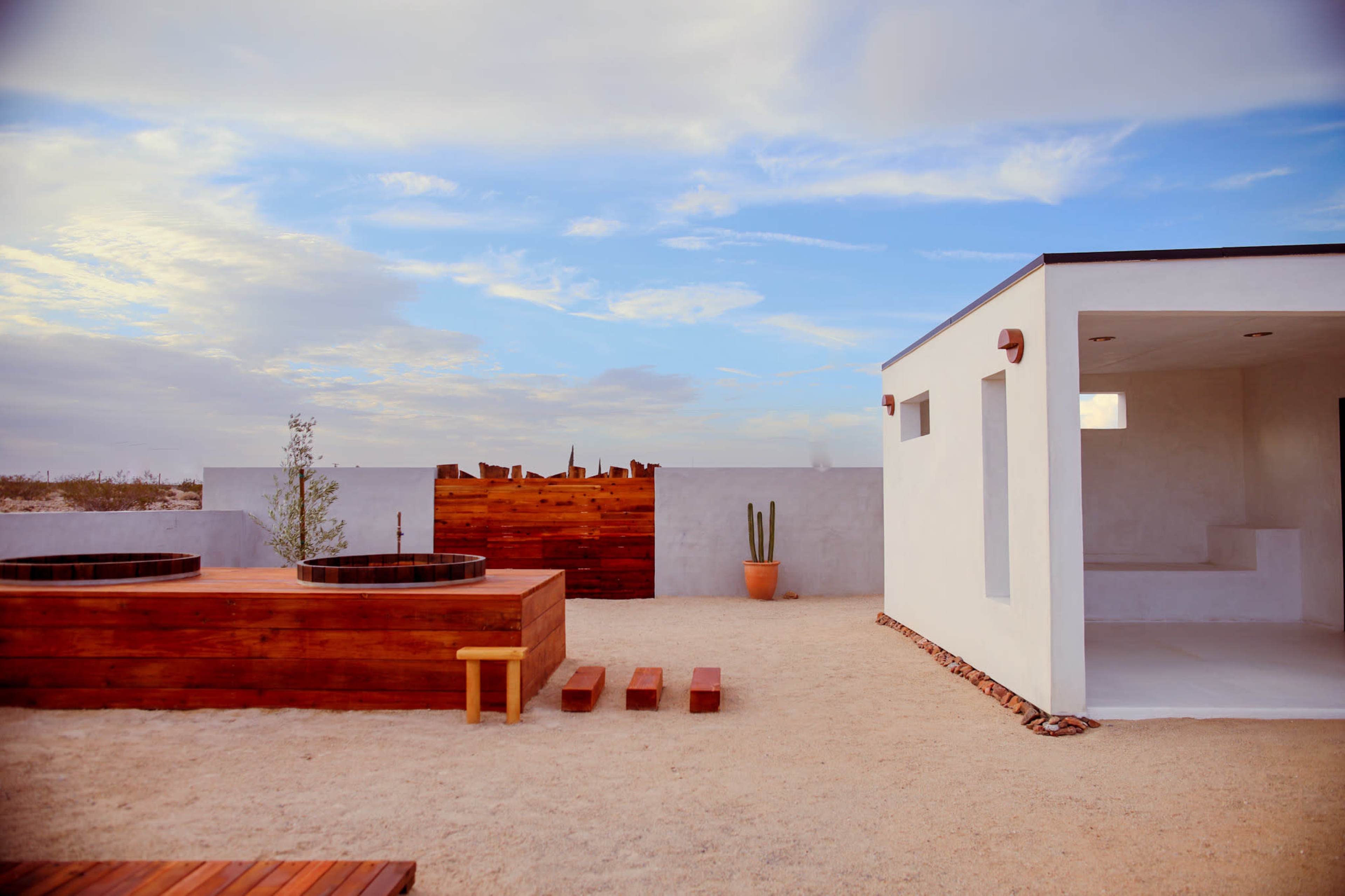 The image shows a modern outdoor space featuring wooden hot tubs, a pathway, a potted cactus, and a minimalistic white building against a cloudy sky.
