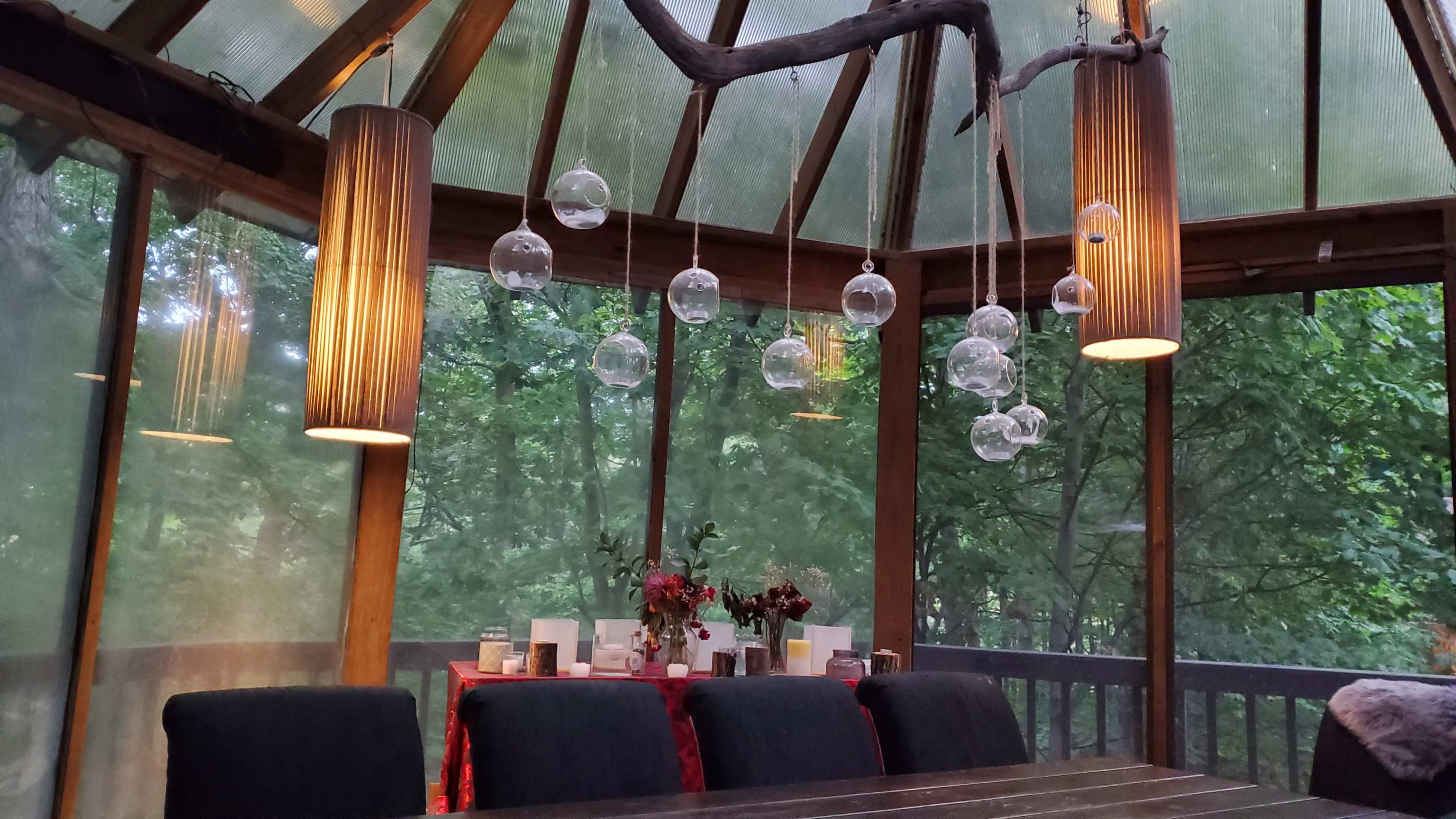 A rustic dining area with a wooden table surrounded by chairs, illuminated by hanging glass globes and cylindrical light fixtures, with greenery visible through the glass walls.