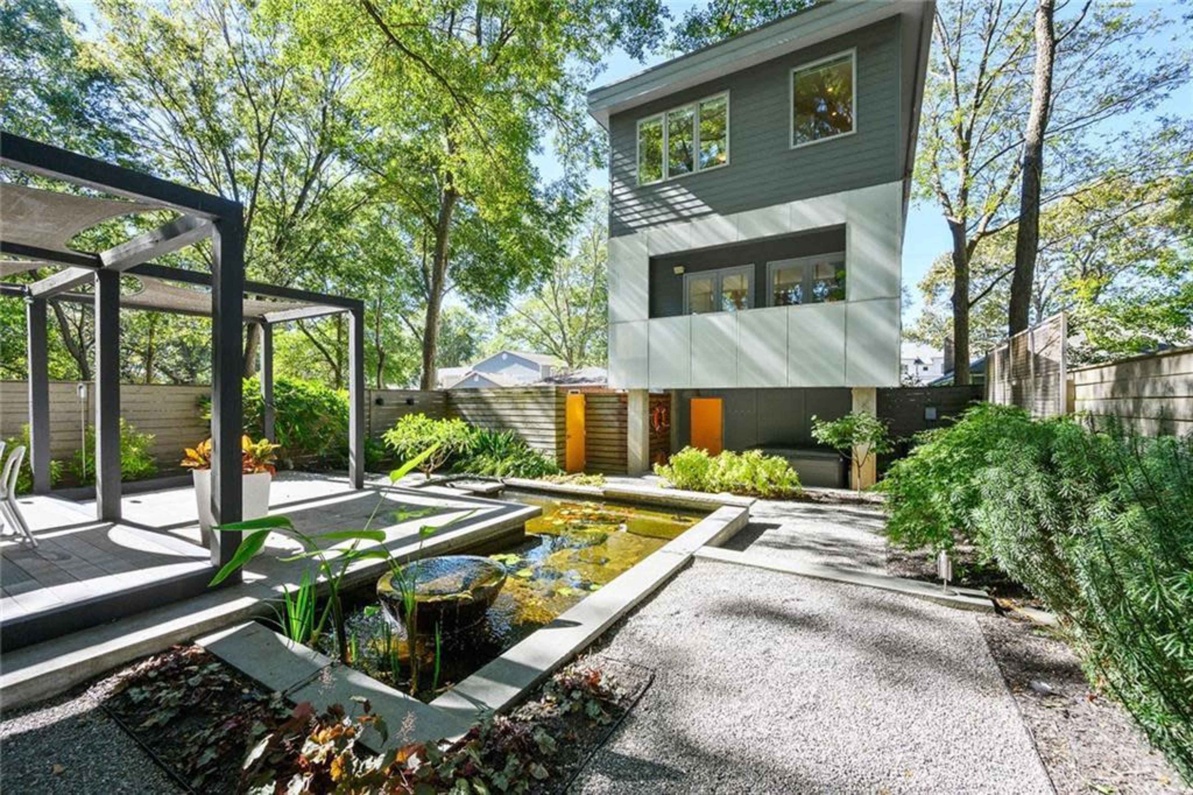 A modern three-story house with large windows overlooks a landscaped garden featuring a pond and stone pathways.