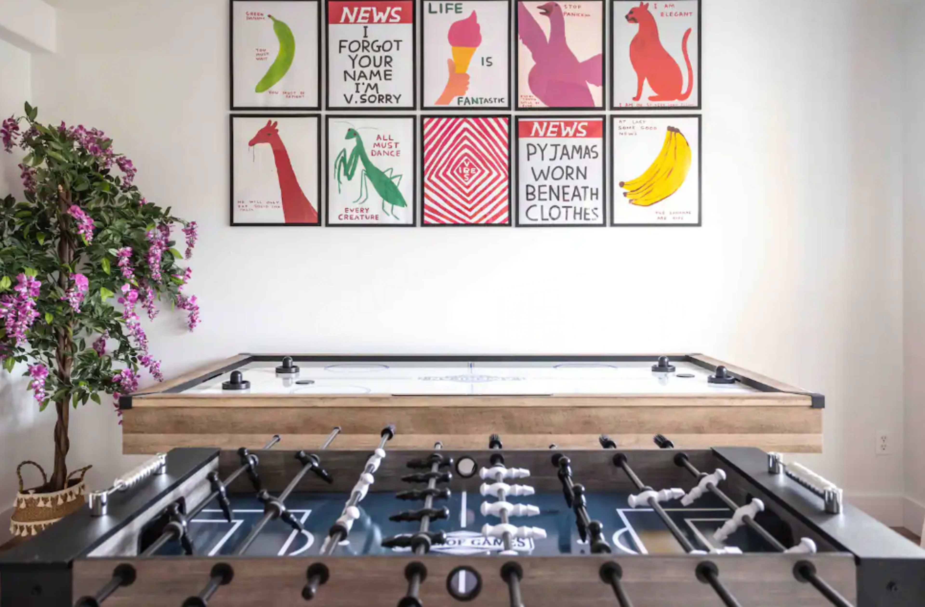 The image shows a game room featuring a foosball table in the foreground and an air hockey table in the background, with colorful framed artwork on the wall above.