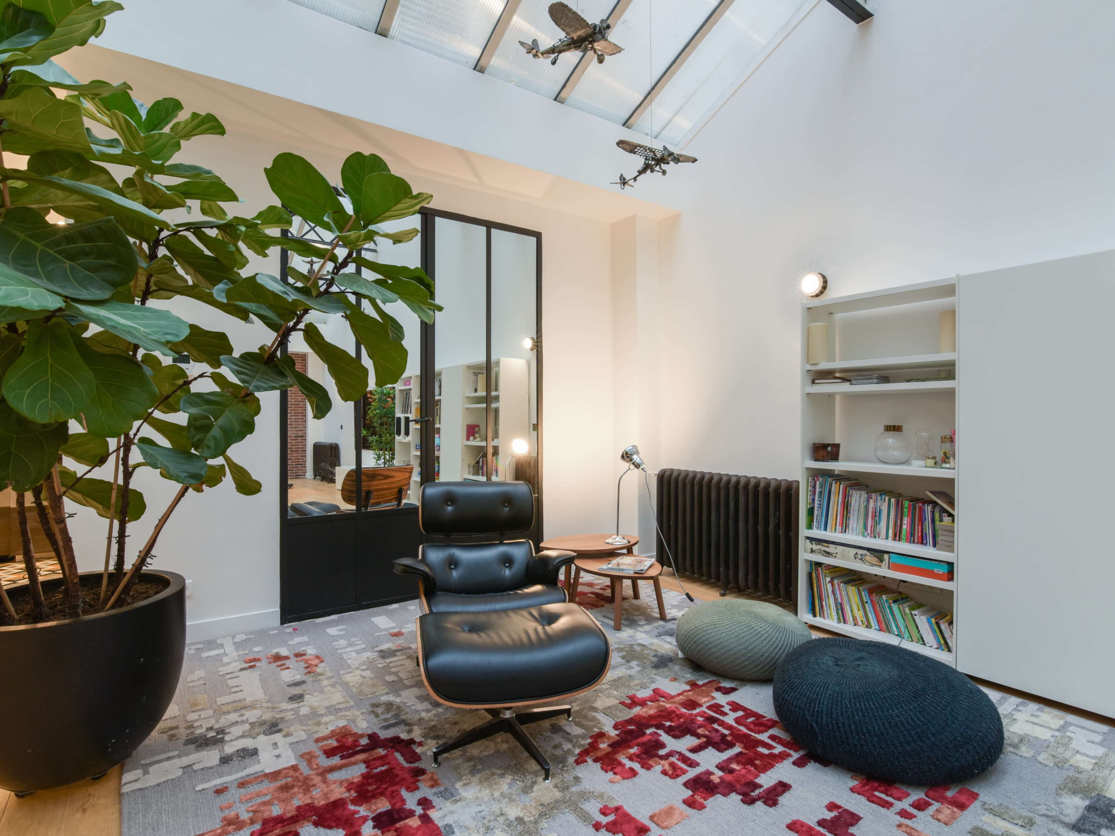 The image shows a modern living space featuring a large plant, an armchair, a bookshelf with various books, and a mix of rugs on the wooden floor.
